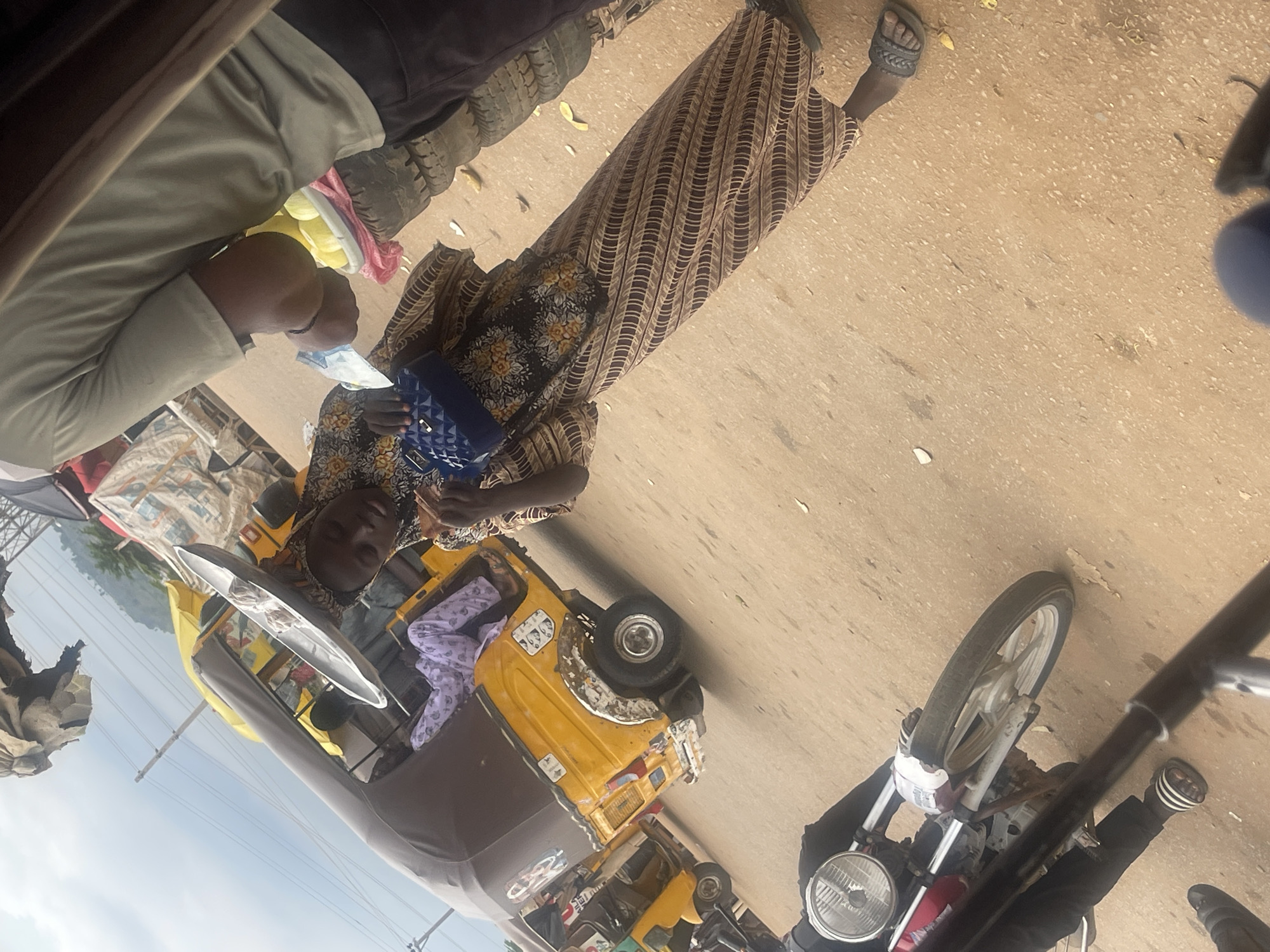 In Galadimawa Village, Nigeria, a woman stands beside a yellow auto-rickshaw. She is wearing a patterned dress and holding a blue purse. Another person, wearing a light-colored t-shirt and dark pants, stands to her right, holding a bowl of what appear to be fruits, possibly mangoes, which are stacked on tires. The background shows more auto-rickshaws, overhead wires, and a cloudy sky. In the foreground, a motorcycle is parked, with its front wheel and handlebars visible. The ground is a dirt road.