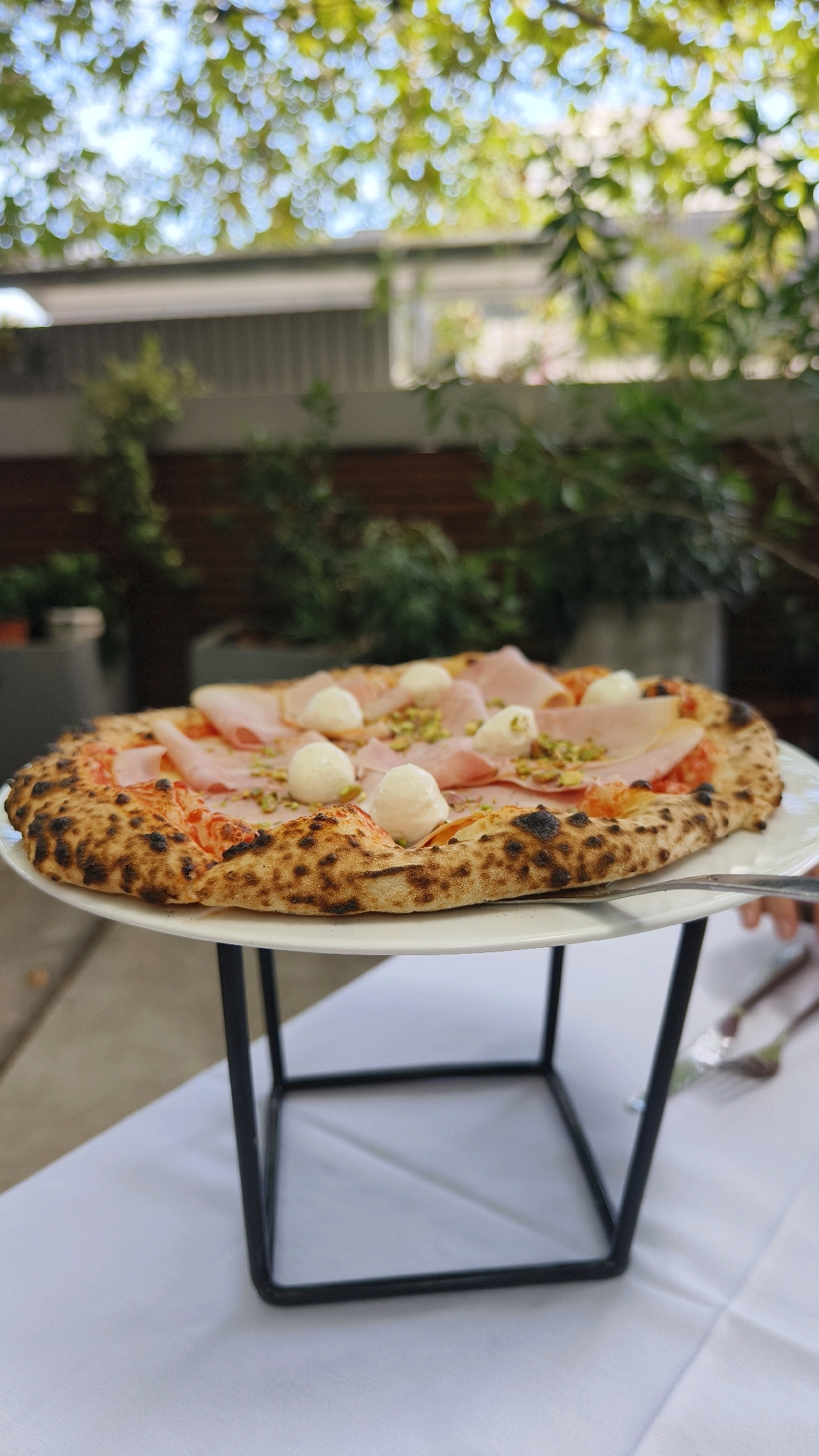 A close-up shot focuses on a pizza served on a white plate, which is elevated on a black metal stand. The pizza features a golden-brown crust with charred spots, topped with thinly sliced ham, small white balls of mozzarella, and chopped green pistachios. The background is blurred, showing greenery from trees and foliage, suggesting an outdoor or garden setting. The scene is lit with natural light, casting soft shadows. The table is covered with a white tablecloth, and a fork is partially visible on the right side of the image, indicating a dining context.