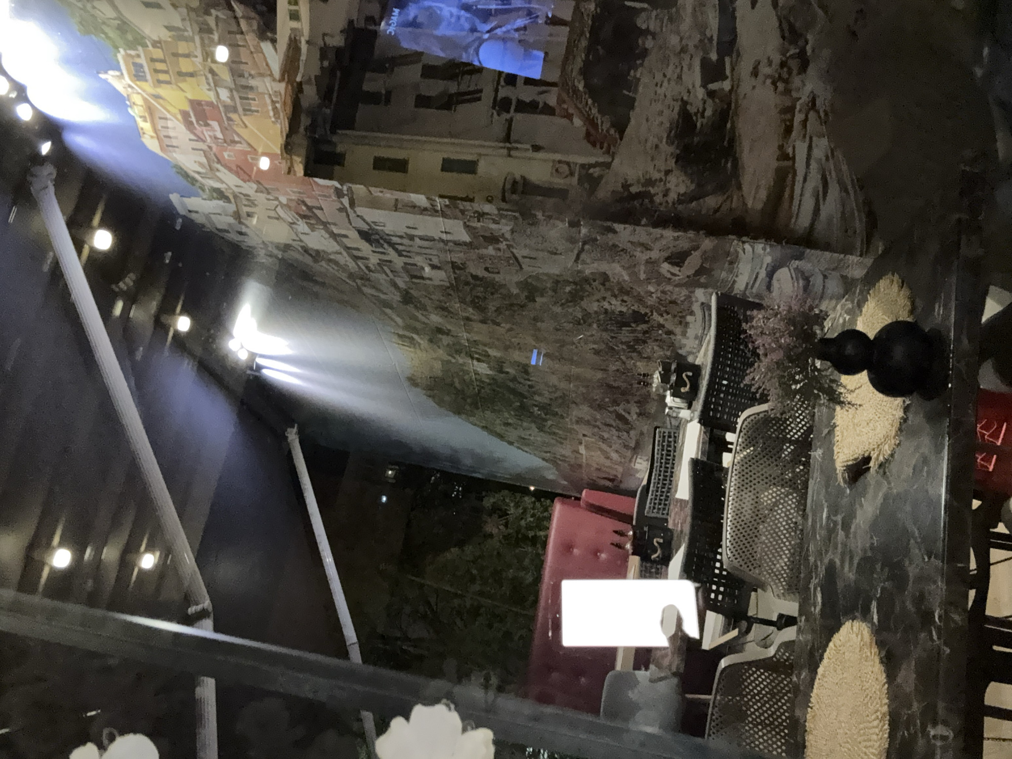 A horizontally-oriented image depicts an outdoor dining area, likely in the city of Sofia, Bulgaria, based on stylistic cues and assumptions. The space is decorated with a large mural depicting a cityscape, possibly of an Italian coastal town. Lighting suggests nighttime. A partial view of structural beams and supports forms a framing element. The dining area features a marble-patterned table with two circular woven placemats. Dark, metal-framed chairs with perforated seats are stacked next to the table. A white rectangular object, perhaps a menu or a napkin, is placed on the table. A red cushioned seating element is also visible. A black decorative object, possibly a set of spheres, is on the table. In the background, beyond the dining area, is foliage, indicating a garden or green space. The overall scene suggests a relaxed and stylish outdoor dining environment.