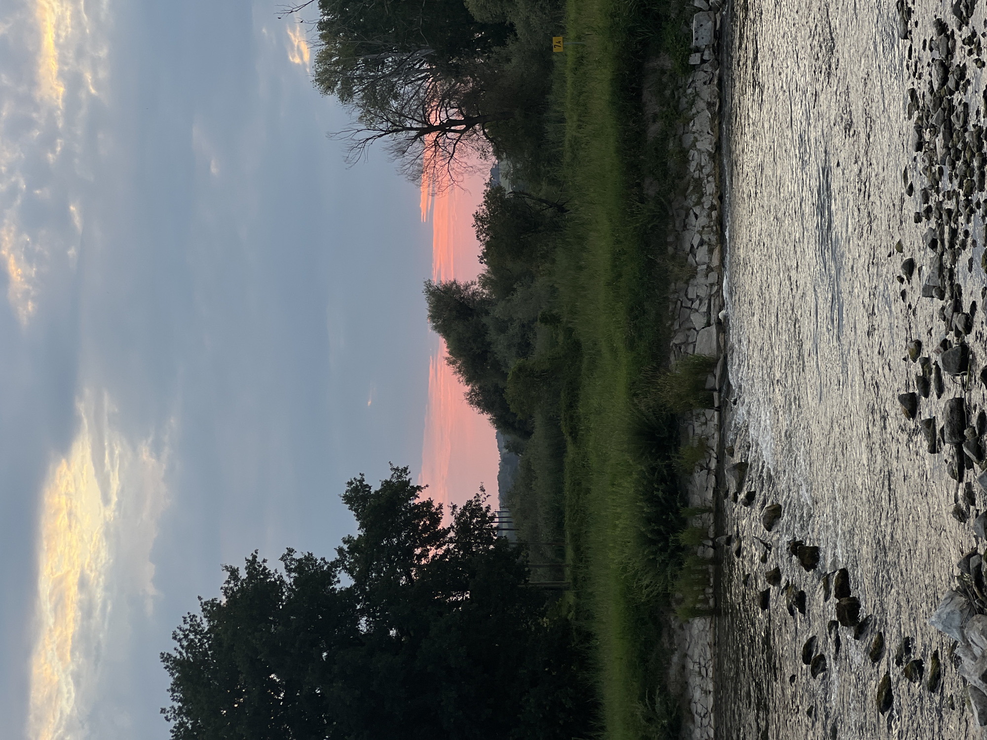 The image depicts a serene river scene at dusk, with the soft hues of a sunset painting the sky. The foreground is dominated by the gently flowing water of the Isar River, scattered with numerous rocks, some submerged and others forming a shallow crossing. The riverbanks are lined with stones and a dense growth of green grass and reeds. Further back, a line of trees and lush vegetation forms a natural barrier, with a large, dark tree silhouette on the left and a more sparse collection of trees on the right. The sky above is a mix of soft gray clouds interspersed with patches of bright, warm light, indicative of the setting sun. A small yellow sign with "7½" is visible on the right bank, suggesting a navigational marker or a distance indicator. The overall atmosphere is tranquil and natural. The location is the Isar River in Moosburg, Germany, specifically the area near the Untere Isarbrücke.