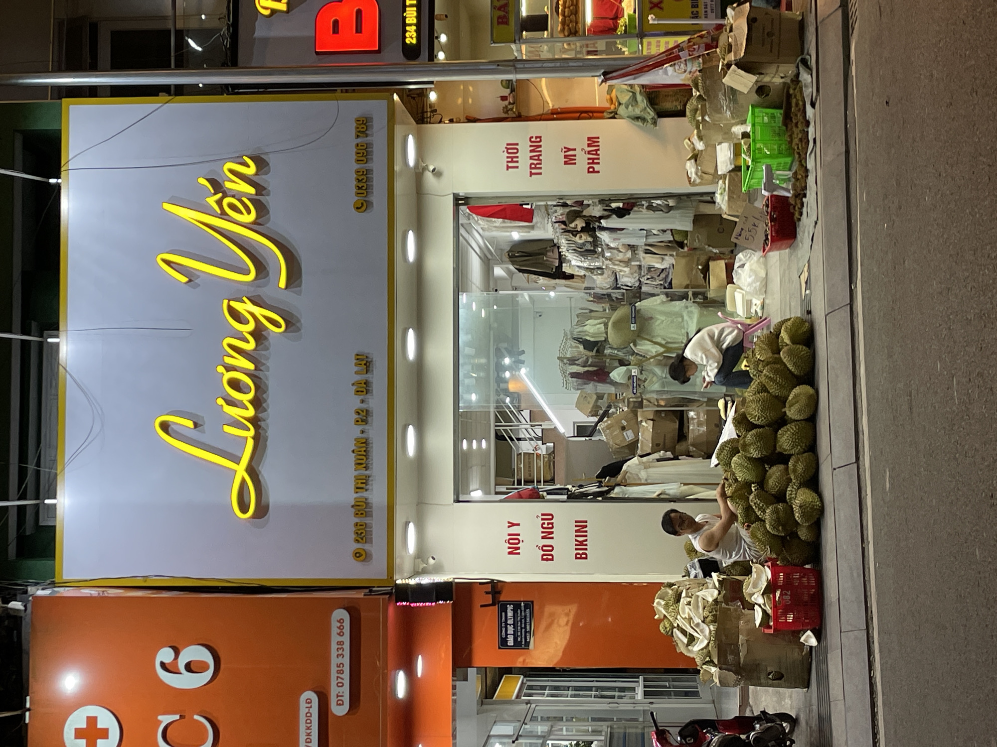 The image captures a street-level view of a commercial establishment in Phường Xuân Hương - Đà Lạt, Vietnam, at night. The establishment features a prominent yellow neon sign with Vietnamese script that reads "Luong Yen." To the left of this sign, an orange facade displays a red cross symbol and white numbers "C 6." Below this, a white oval plaque reads "DT: 0785 338 666."
The storefront itself is a glass-fronted shop displaying clothing and what appear to be mannequins. Text on the white pillar to the right of the entrance lists "NỘI Y," "ĐỒ NGỦ," and "BIKINI" in red, and "THỜI TRANG," "MỸ PHẨM" also in red.
In front of the shop, on the sidewalk and street, a large quantity of durians are piled up. Two individuals are present: one man in a white tank top and glasses sits amongst the durians, appearing to be sorting or preparing them. Another person, possibly a woman wearing a white sweatshirt, is seated on a pink stool to the right, looking down at something in her lap. Red baskets, green crates, and cardboard boxes containing durians or other goods are also visible. The street is paved, and a portion of a red scooter is visible on the far left. The overall scene suggests a street market or roadside stall operating in conjunction with the shop.