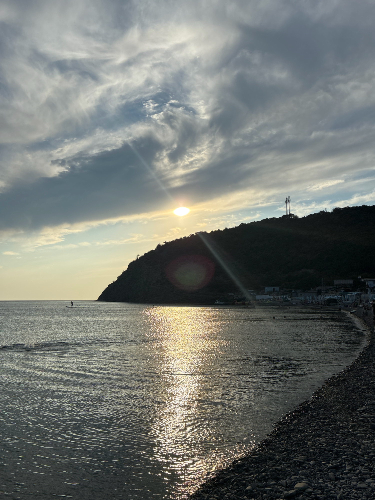 The image depicts a coastal scene at dusk in Dyurso, Russia. The sun is setting, casting a golden shimmer on the calm sea. A silhouette of a person on a paddleboard is visible in the distance. The shoreline is lined with pebbles, and beyond that, a dark, tree-covered hill dominates the landscape. A communication tower stands atop the hill. Several small buildings, possibly beachside structures or residences, are situated at the base of the hill, with people visible along the shore and in the water. The sky is a dramatic mix of dark, heavy clouds and lighter, wispy formations illuminated by the setting sun. The overall mood is serene and tranquil, with a hint of the fading day's warmth.