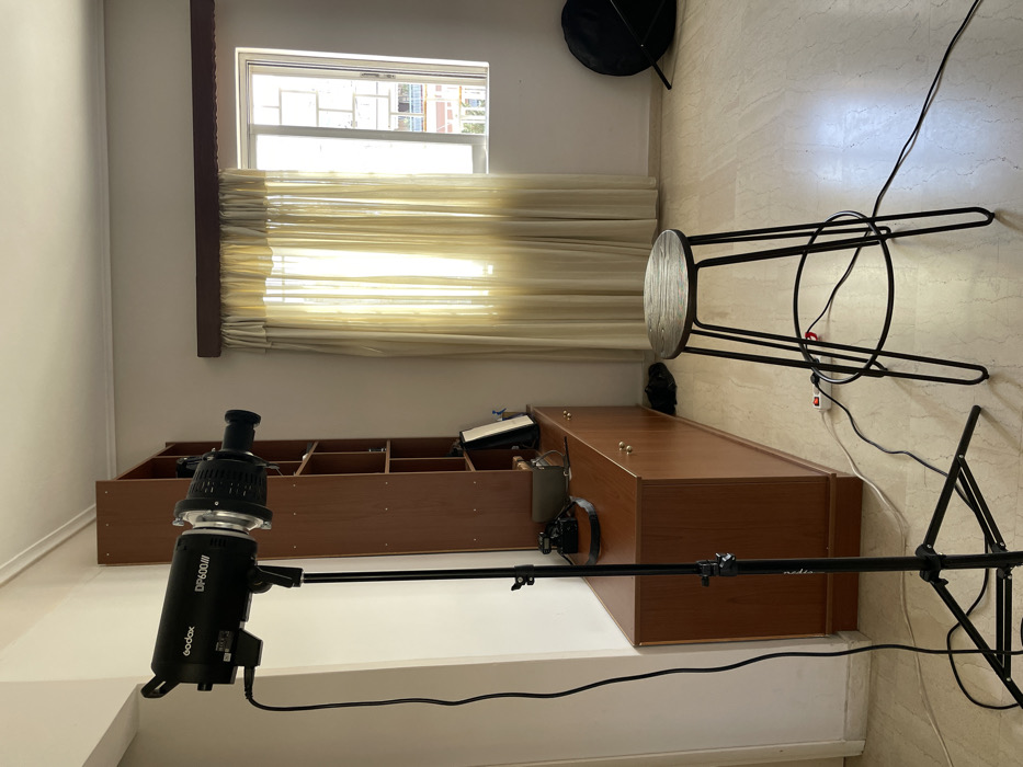 This image captures an indoor room in Vironas, Greece, set up for photography. The primary subjects include professional lighting equipment, furniture, and a window providing natural light.
In the foreground and central to the image, a black Godox DP600III studio flash unit is prominently displayed. It is mounted on a black boom arm, which in turn is supported by a light stand, extending horizontally into the room. The boom arm has "Prolux" text visible. Power cables from the flash run towards a power strip on the floor.
The room features light-colored walls and a light-toned, possibly tiled, floor. Along the back wall, a long, low brown wooden storage unit or bookshelf is visible, filled with various items. Above this unit, a window with light beige horizontal blinds allows strong natural light to stream in, casting bright patterns across the blinds and the wall. Through the upper, uncovered part of the window, residential buildings can be seen, indicating an urban or suburban environment. To the right, a minimalist stool with a black metal frame and a round wooden seat is partially visible, appearing to be stored against the wall.
The scene depicts a functional space, likely a home studio or a room temporarily converted for a photoshoot. The strong sunlight suggests it is daytime. No people are present, so there is no specific activity or event taking place, but the setup clearly indicates preparation for photography.