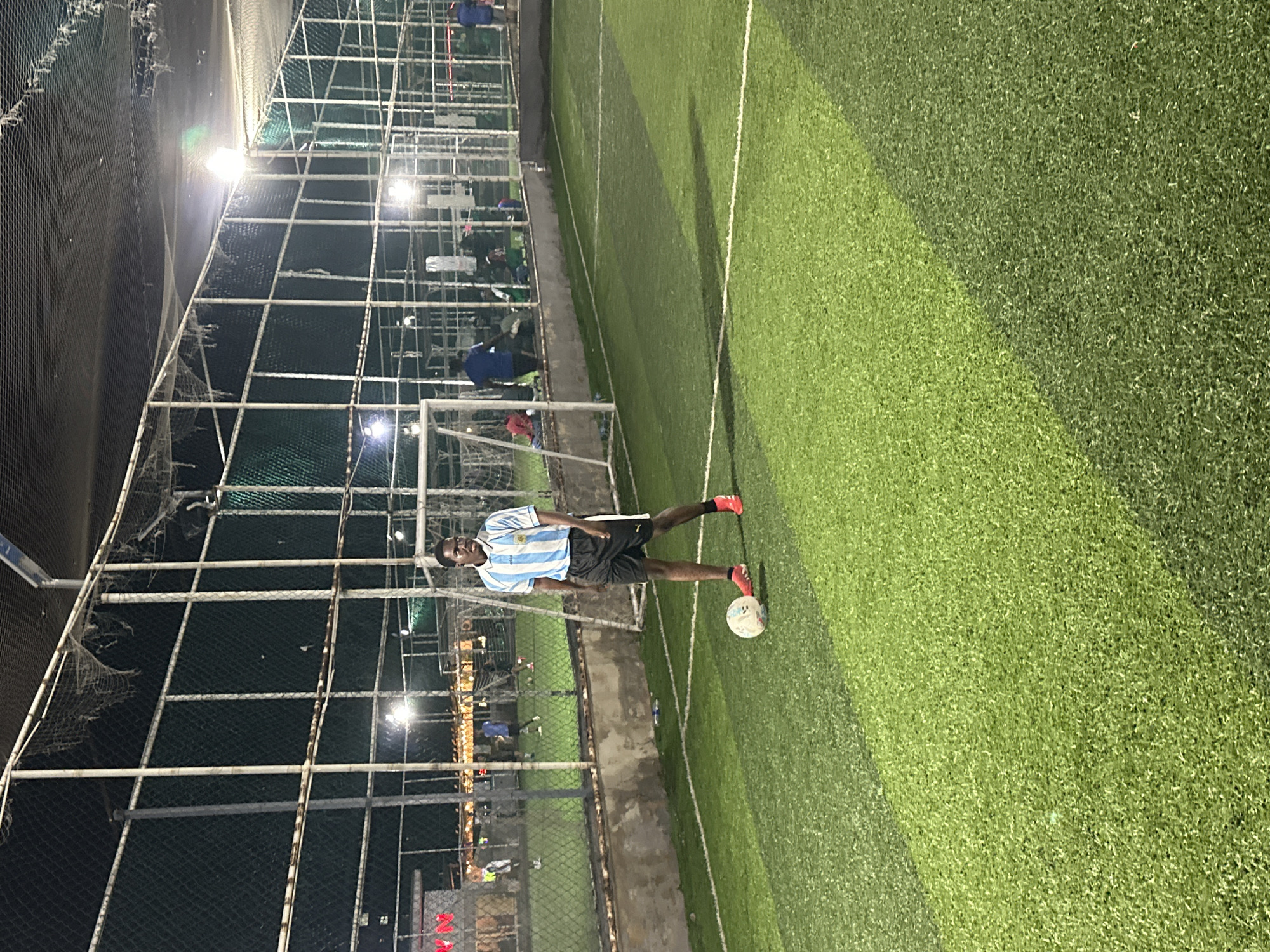 In the city of Abuja, Nigeria, at night, a young Black man stands on an artificial turf soccer field. He is wearing a blue and white striped soccer jersey with white trim and black shorts. His socks are red. He is looking to his left, with his right foot positioned near a white soccer ball. The field is lit by artificial lights, and a chain-link fence with netting is visible in the background. Other people are scattered in the background, some sitting and others standing, near structures that appear to be part of a sports complex. The overall atmosphere suggests an evening practice or game.