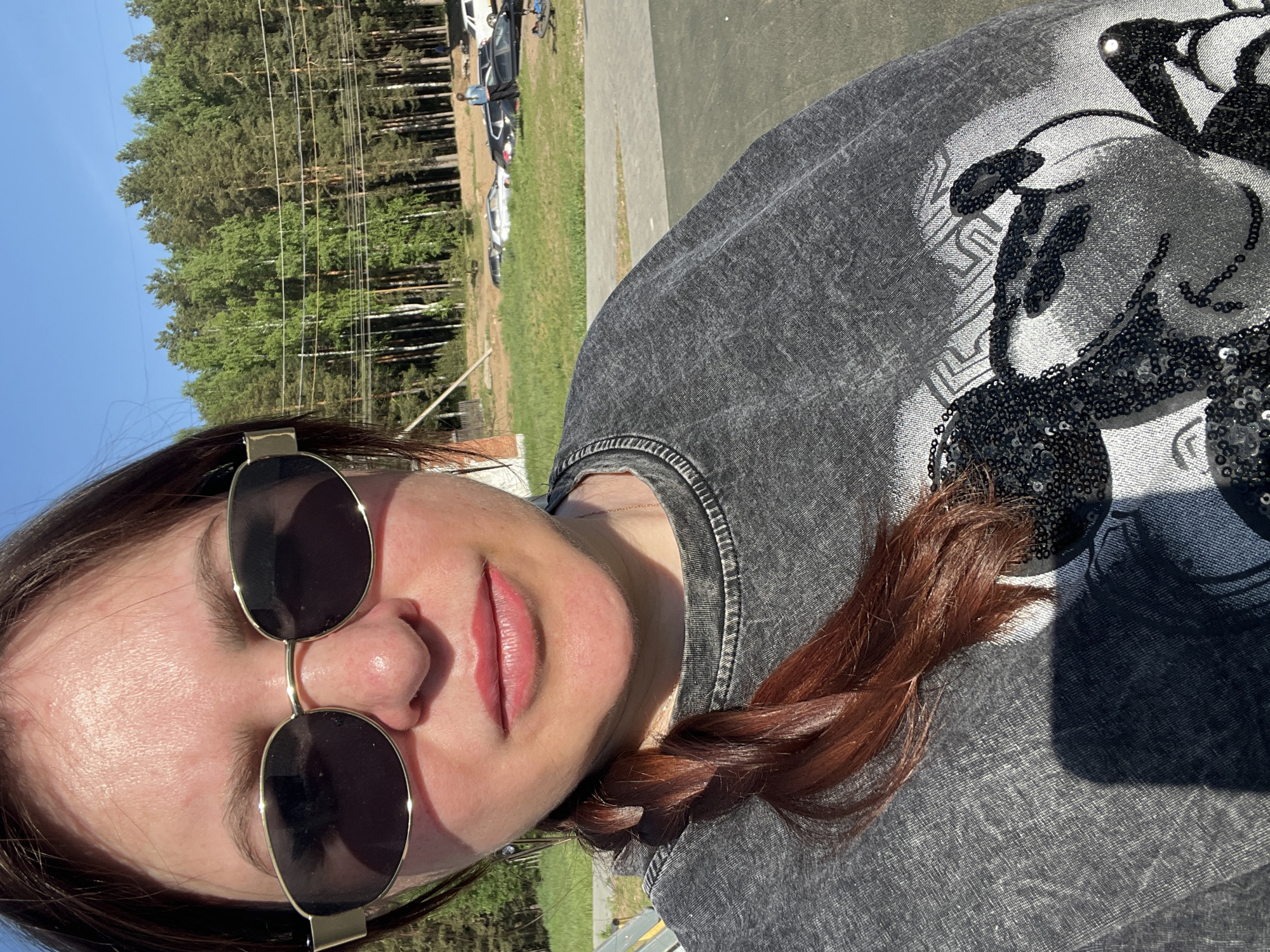 A woman poses in an outdoor environment. She wears sunglasses and a gray shirt with a sequined design. Her hair is braided. The background features trees, grass, and parked cars. Based on the context provided, the location is Noviy, Russia.