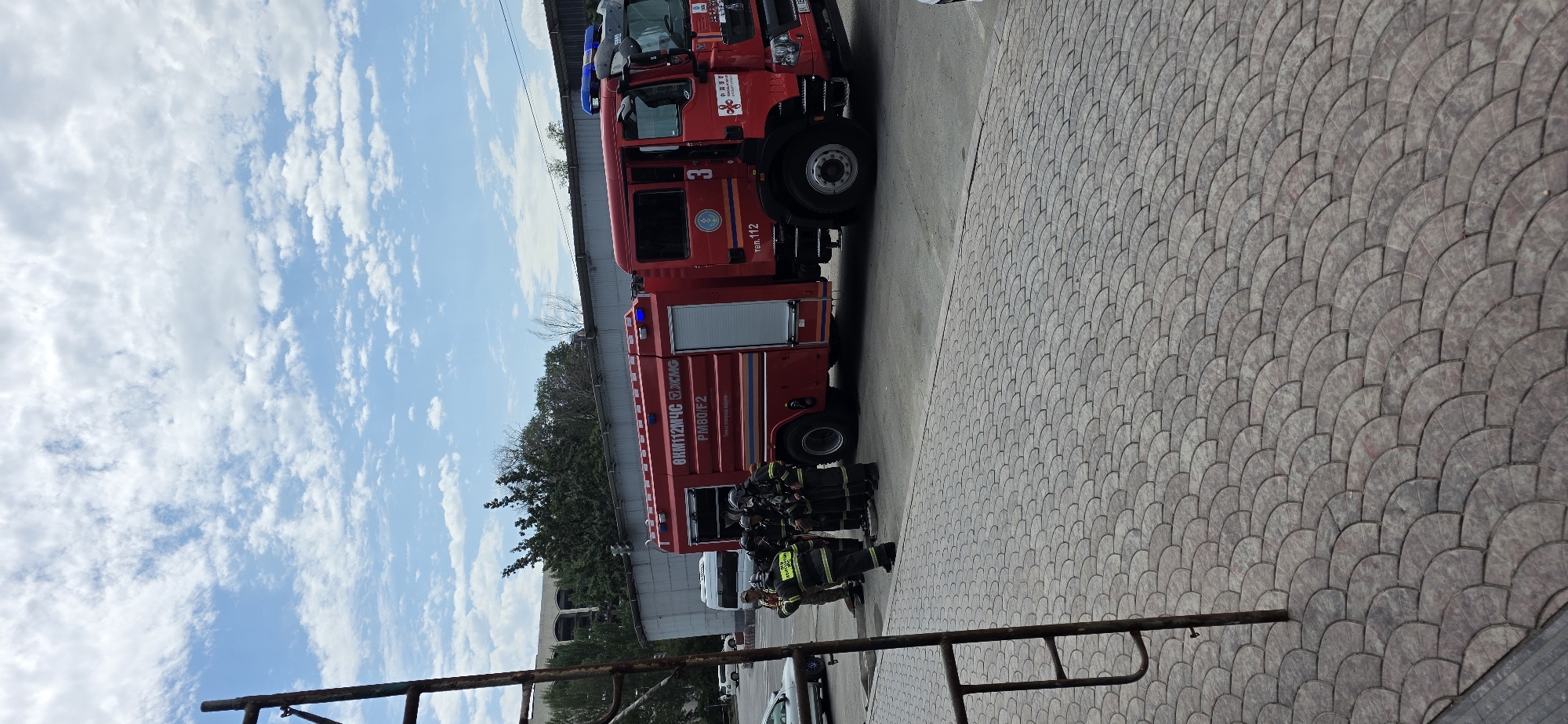 In Bishkek City, Kyrgyzstan, on a cloudy day, a red fire truck with blue lights is parked on a paved surface. Several firefighters in full gear are gathered around the truck, some appearing to be in conversation or receiving instructions. A metal structure, possibly scaffolding, is visible in the foreground on the left. In the background, there are trees, a building with damaged windows, and other vehicles, including a white van and a silver car. The scene suggests a response to an incident, with emergency personnel actively engaged.