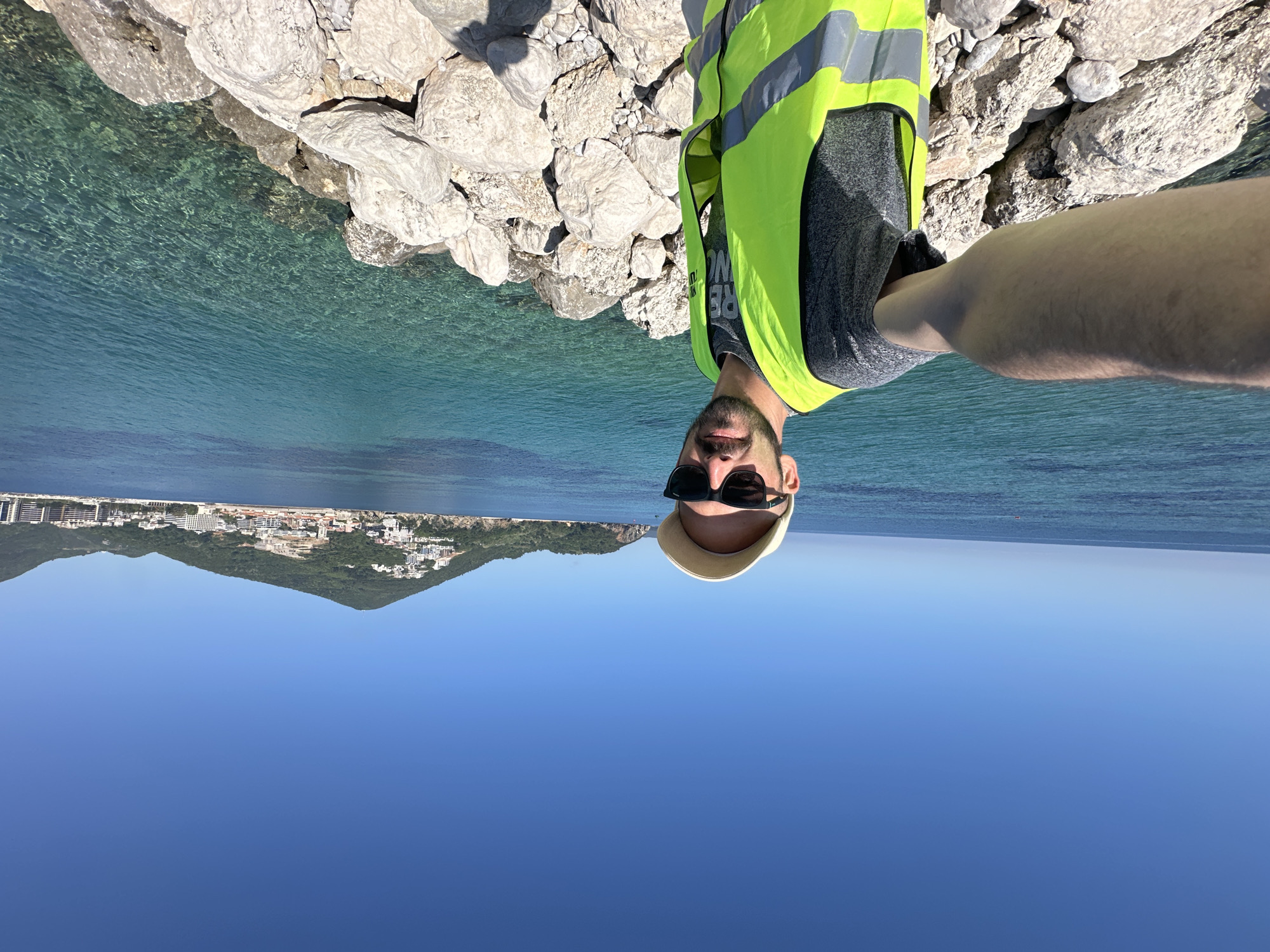 A selfie shows a man wearing sunglasses, a white hat, and a fluorescent yellow vest, standing on a rocky shoreline. The background includes a clear blue sky, deep blue ocean, and in the distance, the coastal city of Budva, Montenegro nestled beneath a mountain. The man's arm extends into the frame to take the photo. The water near the rocks is clear and turquoise, revealing the seabed.