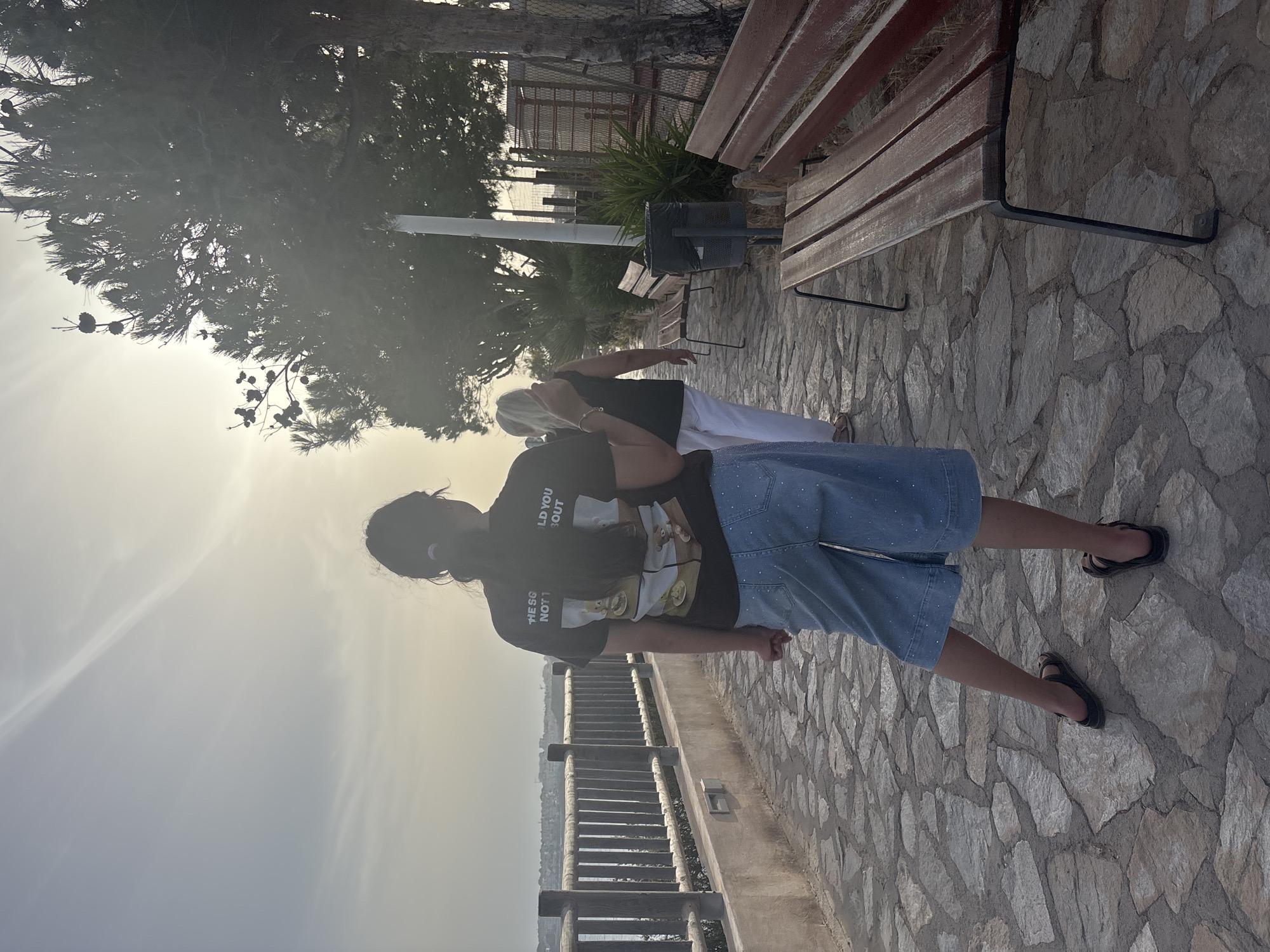 A young girl with dark hair in a ponytail, wearing a black t-shirt and light blue denim shorts with white polka dots, walks along a stone-paved path. She is seen from behind, and her feet are bare in dark sandals. Beside her, slightly ahead, a woman with blonde hair, wearing a black sleeveless top and white trousers, also walks away from the camera. The path is bordered by a wooden railing on the left and benches and trees on the right. In the background, a body of water can be seen with a distant coastline. The scene appears to be a promenade in Orihuela, Spain, possibly near the coast, with the sun setting or rising, casting a warm glow over the landscape and creating lens flare. The sky is a pale yellow and blue. The benches are made of wood with black metal legs, and a dark trash can is situated between two of them. The overall atmosphere is serene and peaceful.