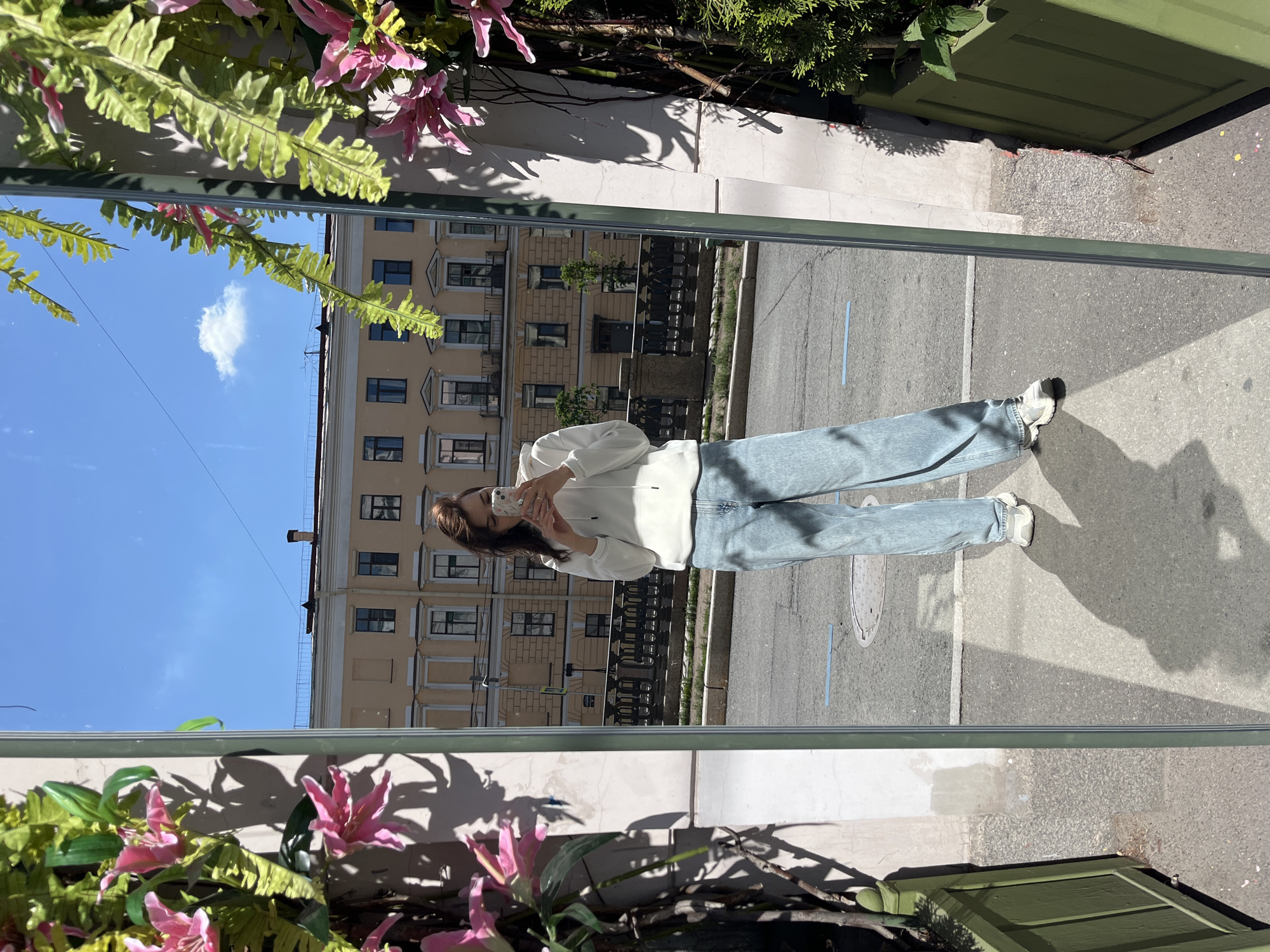 A young woman stands in front of a full-length mirror, taking a selfie. She is wearing a white hooded sweatshirt and light-wash, wide-leg jeans. The mirror reflects a street scene in Saint Petersburg, Russia. Behind her, a building with classical architecture, featuring multiple windows and balconies, lines a canal. The sky is clear and blue with a single white cloud. Green planters with pink flowers and foliage flank the mirror, adding a touch of nature to the urban setting. The woman's shadow is cast on the pavement, indicating a bright, sunny day. The overall impression is one of a casual, everyday moment captured in a picturesque city.