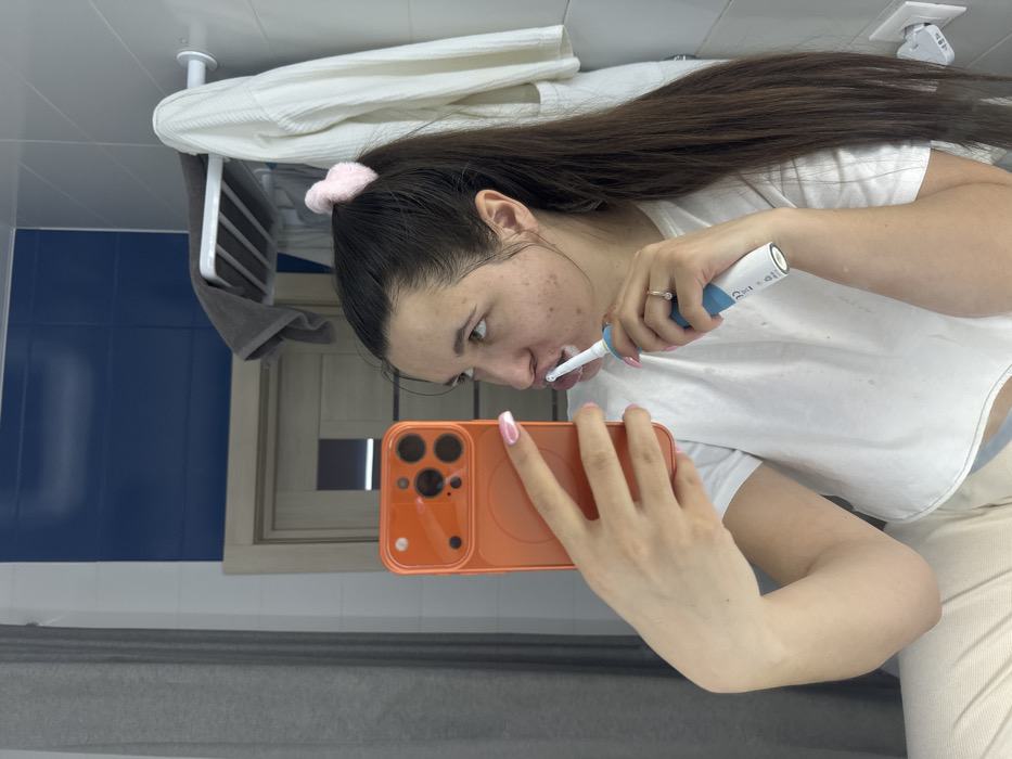 The image depicts a female individual taking a mirror selfie in a residential bathroom, identified as being in the city of Odintsovo, Russia. The subject has long, dark brown hair tied in a high ponytail with a light pink scrunchie. She is wearing a white crop top and light-colored bottoms. Her left hand holds an orange-cased smartphone with a prominent rear camera array, positioned to capture her reflection. Her right hand operates a blue and white electric toothbrush, which is in her mouth with visible toothpaste foam, indicating active tooth brushing. A ring is present on the ring finger of her right hand, and her fingernails display pink nail polish, possibly French tips. The bathroom features white and dark blue tiled walls. In the reflection, a light-colored wooden door with a glass panel is visible in the background. To the right, a white multi-bar towel rack holds a grey towel, and a white robe hangs nearby. A white electrical outlet is visible on the right wall, and a grey curtain or textile hangs on the far left.