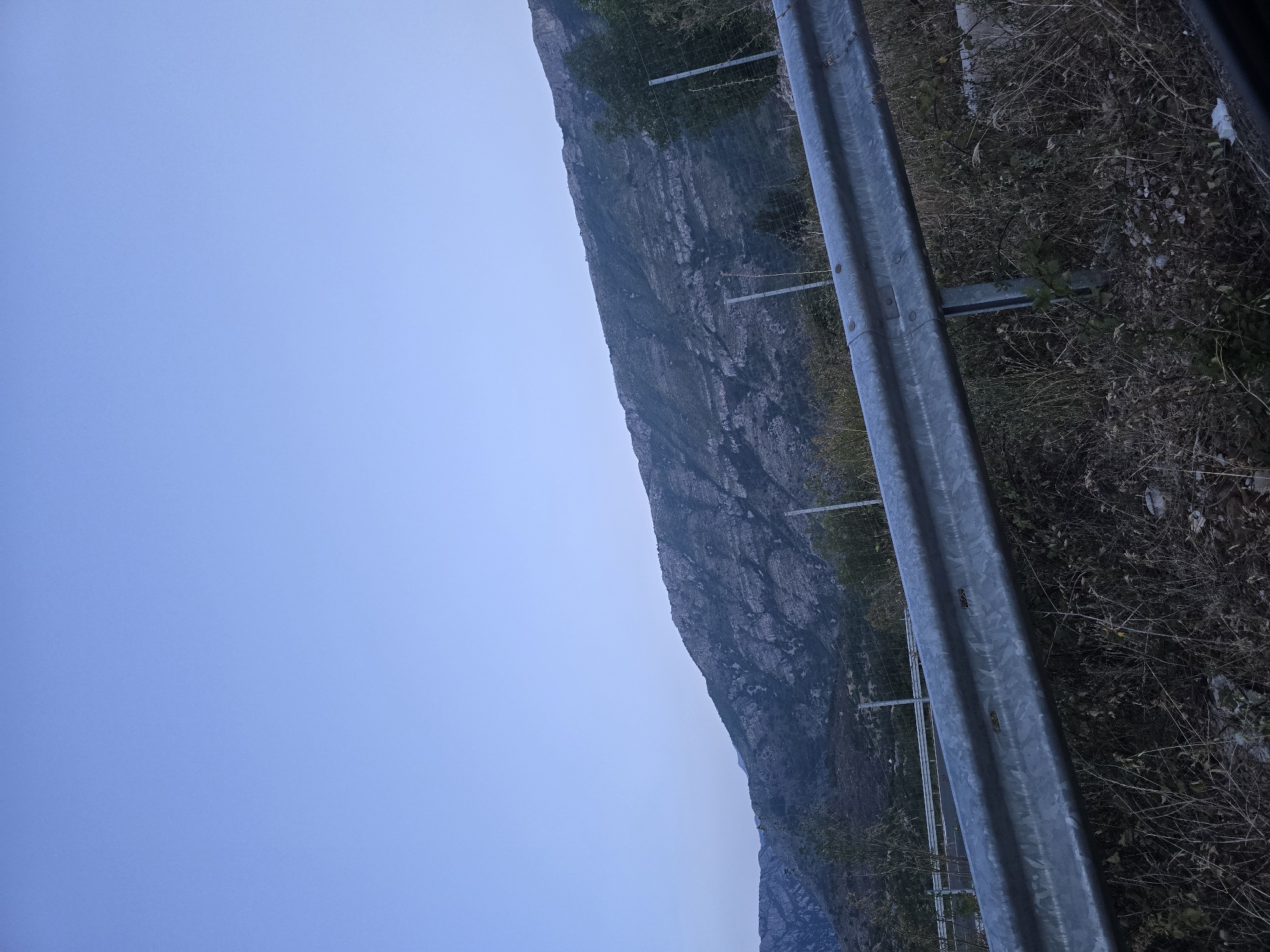 An outdoor shot shows a wide, clear blue sky over a mountain range. The foreground is dominated by a metallic guardrail running diagonally across the frame, with dry grass and foliage beneath it. Behind the guardrail, a fenced area is visible with several posts. In the mid-ground, more vegetation and trees are present, leading up to the rocky, textured slopes of the mountain. The image appears to be taken from a roadside perspective, suggesting a location along a road or highway near Klopot, Montenegro.
