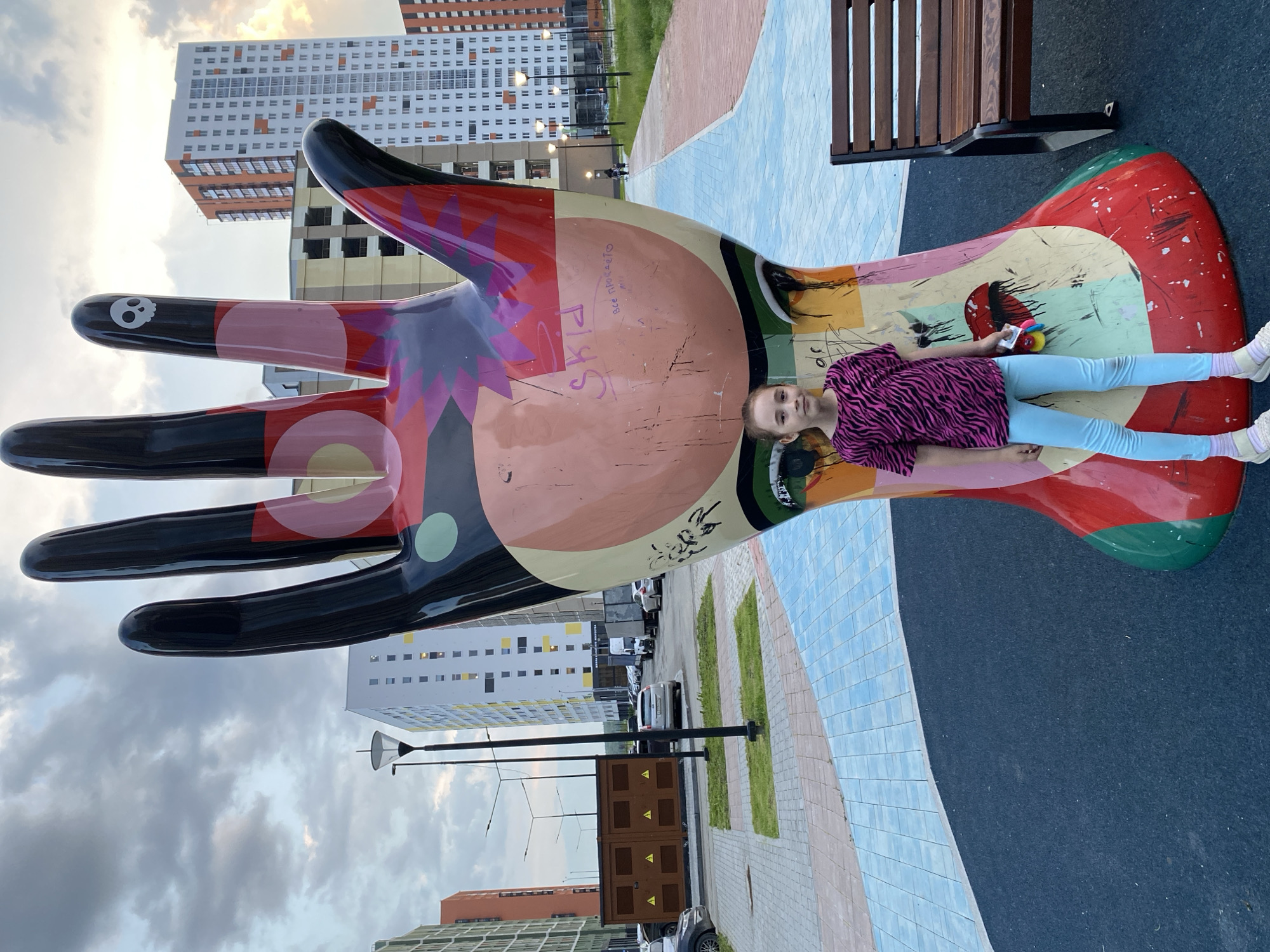 A young girl stands in front of a large, colorful sculpture of a hand in Yekaterinburg, Russia. The sculpture is abstract and painted with various colors and patterns, including pink, red, black, and yellow. The girl, wearing a pink and black striped t-shirt and light blue leggings, holds a toy in her right hand. The background features modern apartment buildings, streetlights, and a paved walkway. The sky is overcast with some visible clouds. The sculpture appears to be a public art installation in a residential area.
