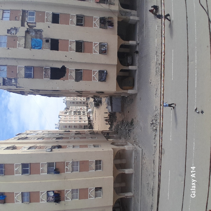 The image, taken with a "Galaxy A14" phone as indicated by the text in the bottom right, captures a street scene flanked by multi-story residential buildings under what appears to be a clear, bright sky with some scattered clouds. The photo is oriented ninety degrees clockwise, depicting the buildings vertically along the left and right sides of the frame, with the street running through the middle.
The most striking feature of the scene is the significant damage sustained by the buildings. The building on the left side of the frame (when viewed upright) shows extensive destruction, with numerous windows either shattered, missing glass panes, or boarded up with dark materials. Several large, irregular holes are visible in its beige-colored facade, exposing dark interiors. One window on this building is covered by a bright blue tarp. The architecture of the buildings features repeating patterns of windows with decorative, possibly arched, frames, separated by vertical strips of a contrasting, slightly darker color. The ground floors of both buildings feature arched open-air walkways or entrances, characteristic of apartment complexes.
In the background, beyond the immediate street and buildings, more multi-story buildings can be seen, also appearing to be part of a densely built-up area. This distant area appears somewhat dusty or desolate, with scattered debris and what might be temporary structures or tents. Piles of rubble and debris are visible on the unpaved ground between the buildings, further reinforcing the impression of a damaged or neglected environment.
A paved road runs between the two main buildings, exhibiting a faded white dividing line. The edges of the road are rough and contain scattered debris. Several individuals are present in the scene. On the road, in the mid-ground, a person wearing light-colored clothing is walking. Further in the foreground on the right side of the road, two smaller figures, possibly children, are visible – one standing, the other crouching. In the distant, unpaved area between the buildings, a group of people are gathered amidst the debris, their specific activities unclear due to the distance.
The overall atmosphere of the image is one of hardship and the aftermath of conflict or severe decay, with daily life seemingly continuing amidst the ruins. The daylight suggests it is daytime.
