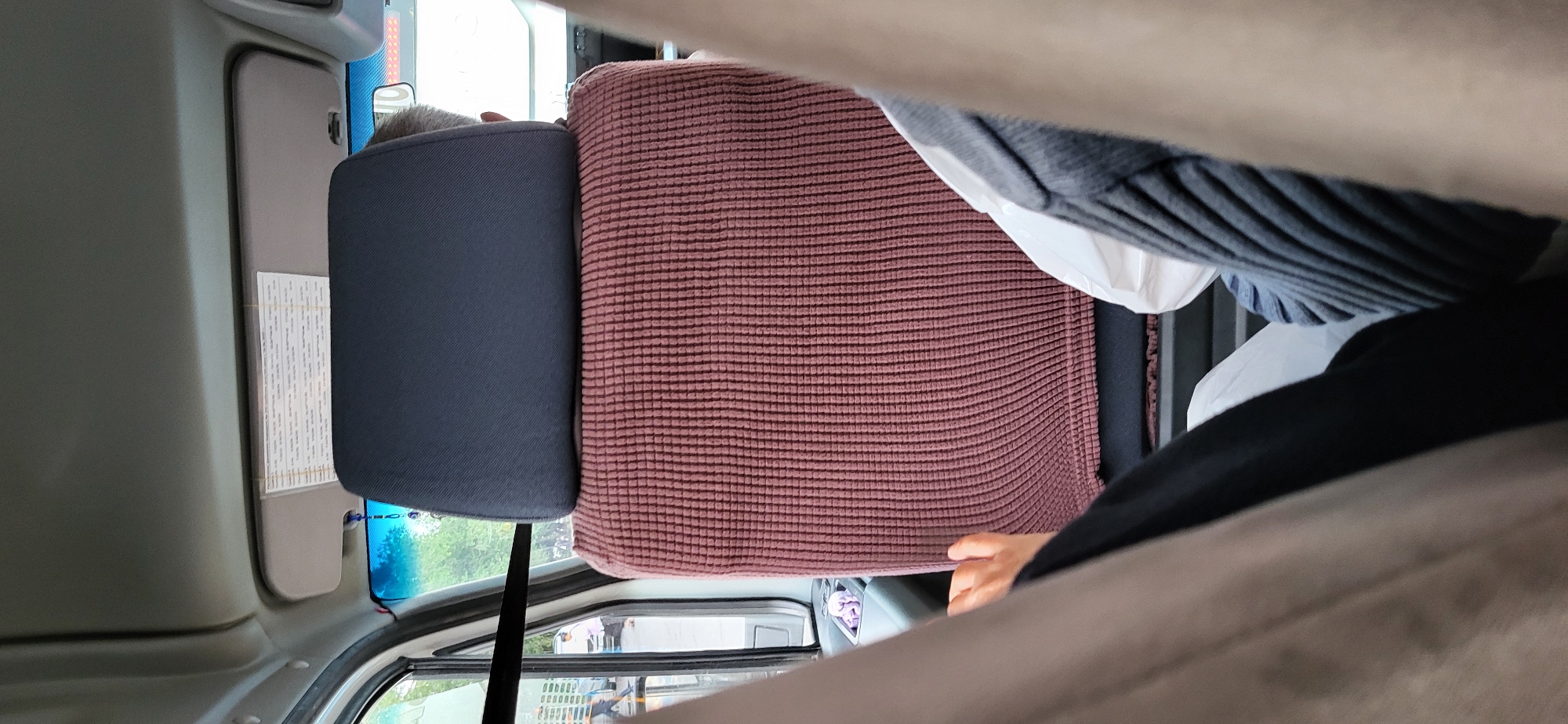 The image shows the interior of a vehicle, likely a van or bus, from a passenger's perspective. The focus is on the back of a headrest and the ribbed fabric seat cover in a reddish-brown color. A person's legs are visible in the foreground, suggesting the photographer is seated behind the driver or in a passenger seat. To the left, through the window, a street scene is visible with people walking and a white vehicle. A sun visor is attached to the roof of the vehicle, with a document or schedule attached to it. The lighting suggests it is daytime. The location is specified as Oskemen, Kazakhstan.