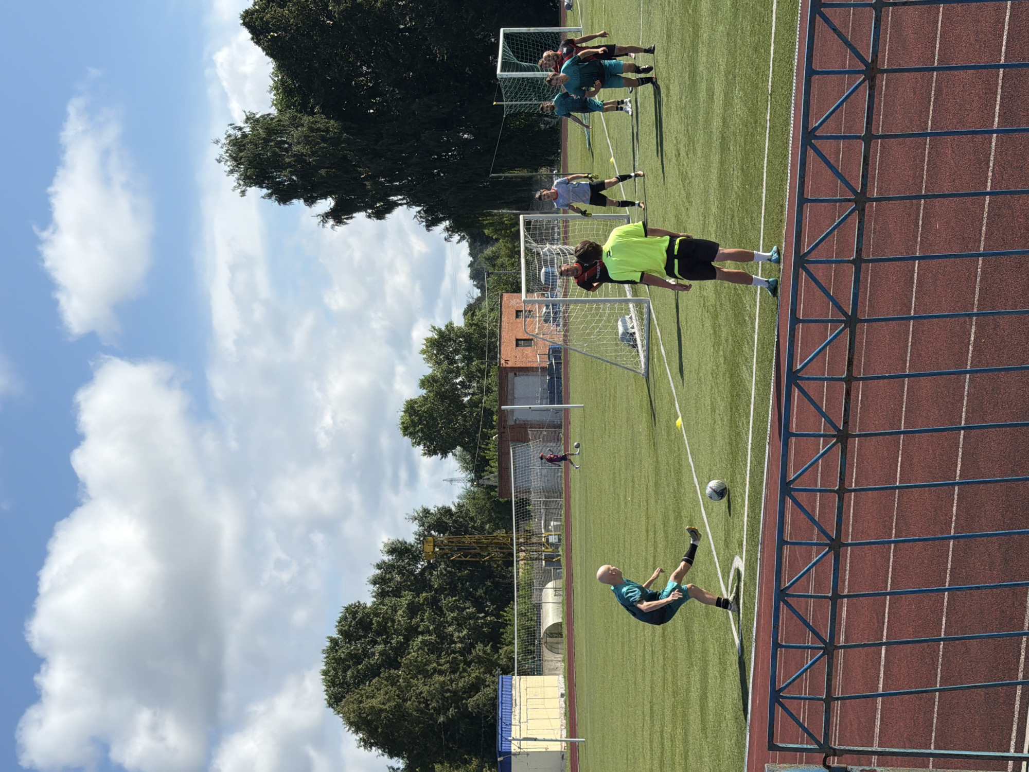 A group of men are playing soccer on an artificial turf field. The field is marked with white lines and has two goals set up. There is a fence in the foreground. In the background, there are trees, buildings, and a stadium light. The sky is blue with white clouds. The image appears to have been taken in the city of Chaikovsky, Russia.