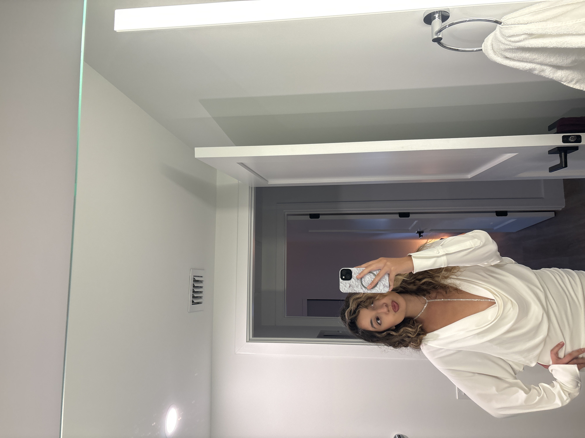 A young woman with curly brown hair takes a mirror selfie in what appears to be a bathroom. She is wearing a white, cowl-neck, long-sleeved dress and a diamond necklace. She is holding a white phone with a marble-patterned case in her right hand. Her left hand is on her hip. The background shows a door ajar, a towel rack with a white towel, and a vent on the ceiling. The scene is set in Hallandale Beach, United States of America.