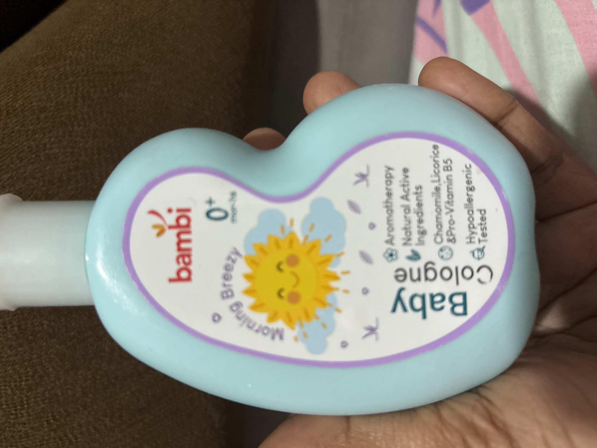 A light blue, ergonomically shaped bottle of Bambi Baby Cologne is held in a person's left hand. The bottle has a white cap and features a purple circular label with a cartoon sun and clouds. Text on the label reads "bambi", "Morning Breezy", "0+ months", "Baby Cologne", "Aromatherapy", "Natural Active Ingredients", "Chamomile, Licorice & Pro-Vitamin B5", and "Hypoallergenic Tested". The background is a brown textured surface and a pink and white striped fabric. The image appears to be taken indoors. The location of Geulanggang Teungoh, Indonesia is not determinable from the image.