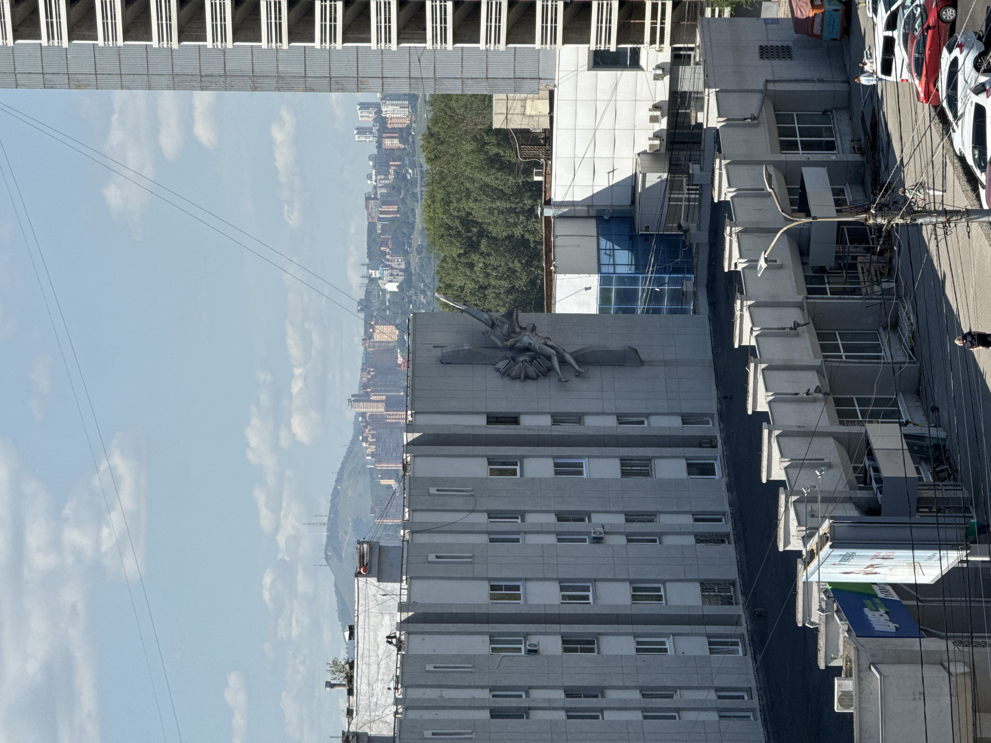 The image captures a cityscape on a bright, sunny day, likely in Krasnoyarsk, Russia, given the architecture. In the foreground, a low-rise building complex with a series of angular, concrete facades and protruding sections is visible. Several large billboards, one with blue and green lettering and another with an image, are positioned near this building. A street with parked cars, including a white SUV, a red sedan, and a white hatchback, stretches in front of the complex. Electrical poles with numerous wires crisscross the scene. Two individuals are visible on the street, one in a dark dress and another in green attire, seemingly walking.
Behind the foreground structures, a mid-rise apartment building with a patterned concrete exterior and numerous windows is prominent. A distinctive sculptural element, appearing to depict figures or drapery, is attached to its facade. Further in the background, a dense line of green trees forms a natural barrier. Beyond the trees, a city skyline is spread out, characterized by numerous buildings of varying heights, including residential blocks and taller structures. In the distance, a hazy, green mountain range with several radio towers on its peak dominates the horizon under a pale blue sky with scattered white clouds. To the right of the mid-rise building, the unfinished concrete structure of a high-rise building is visible, with multiple floors of balconies. A small red and orange vehicle is parked to the far right.