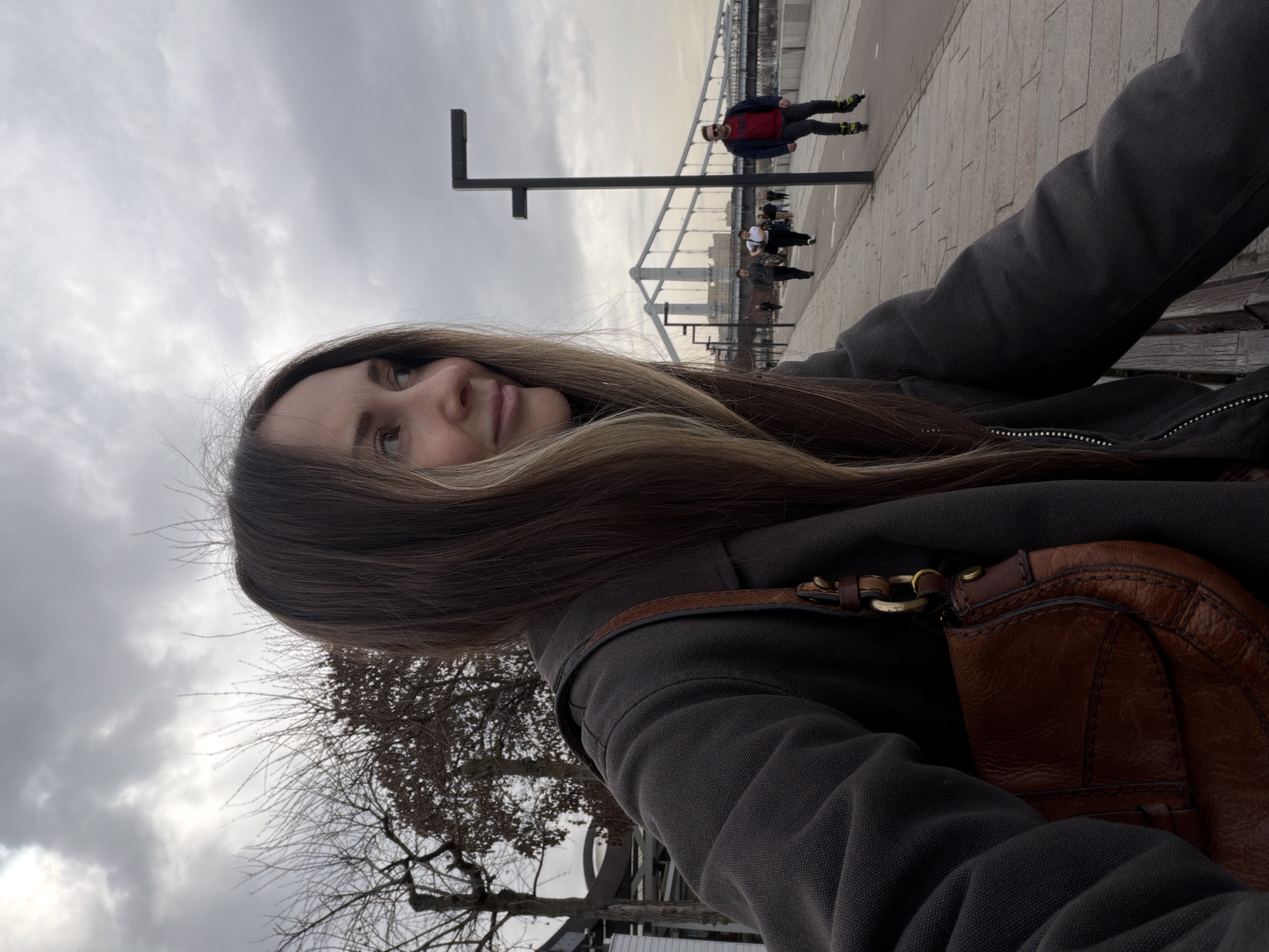 The image captures a selfie-style shot of a person with long brown hair and fair skin, positioned at an angle to the camera. The person is wearing a dark gray jacket and a brown leather handbag with visible metal hardware.
In the background, there is a city scene under a cloudy sky. A pedestrian walkway is visible, populated by several individuals, including one person on rollerblades wearing a red jacket. A bridge structure with metal railings is also present in the distance.
The foreground includes a tree with bare branches and urban elements like a street light pole. The location is the city of Moscow, Russia.