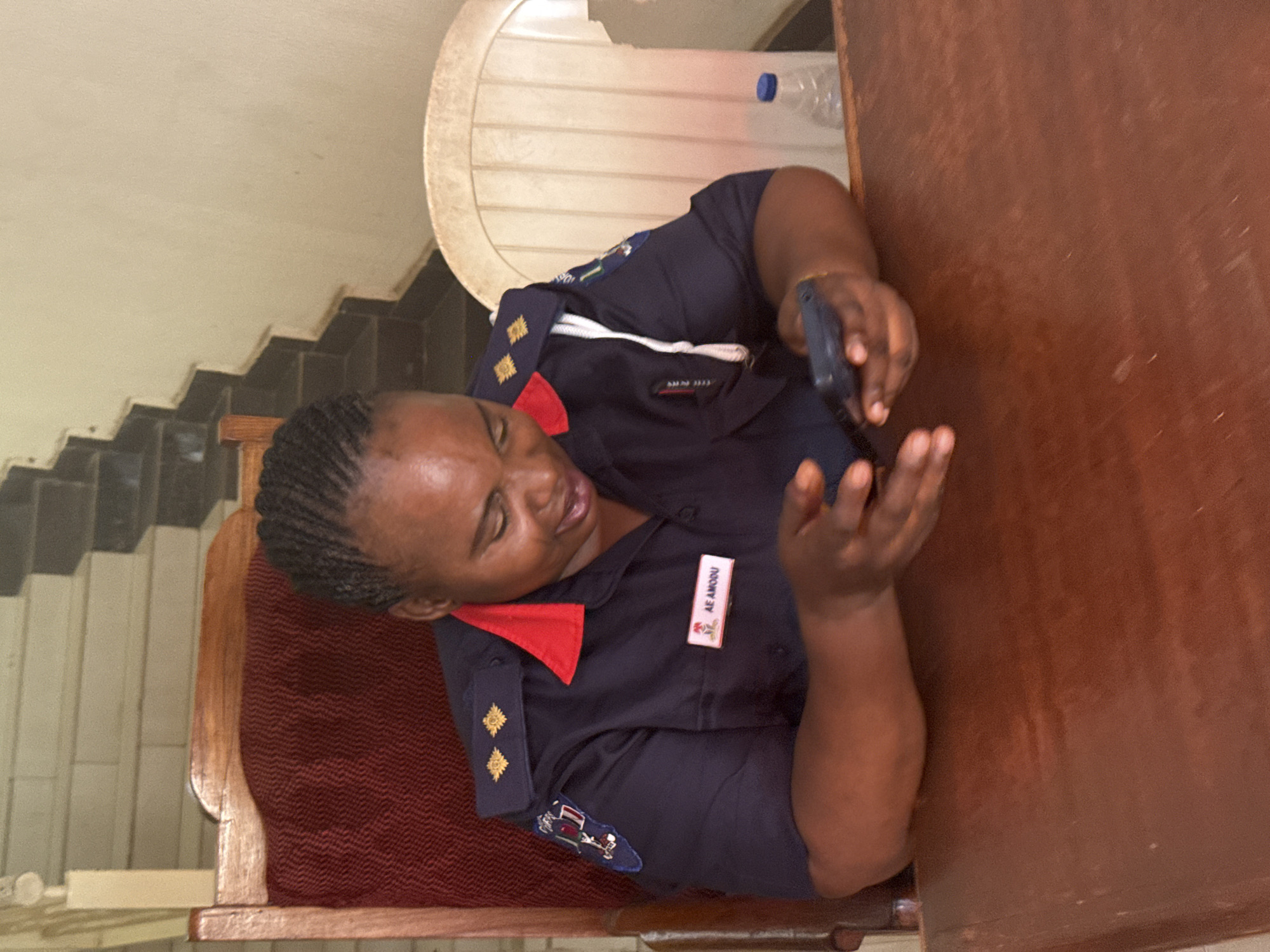 A woman wearing a dark blue uniform with a red collar and embroidered shoulder patches sits at a wooden table, looking at a black mobile phone in her hands. Her uniform has a name tag reading "AE AMOSU" on the right side and two gold badges on each shoulder epaulette. She is seated on a wooden chair with a maroon cushion. A white plastic chair is partially visible behind her, with a plastic water bottle on its seat. To the left are light-colored steps or tiled flooring. The table is large and appears to be made of dark wood. The location appears to be indoors, but the specific details to confirm that the location is the city of Abuja, Nigeria are not present in the image.
