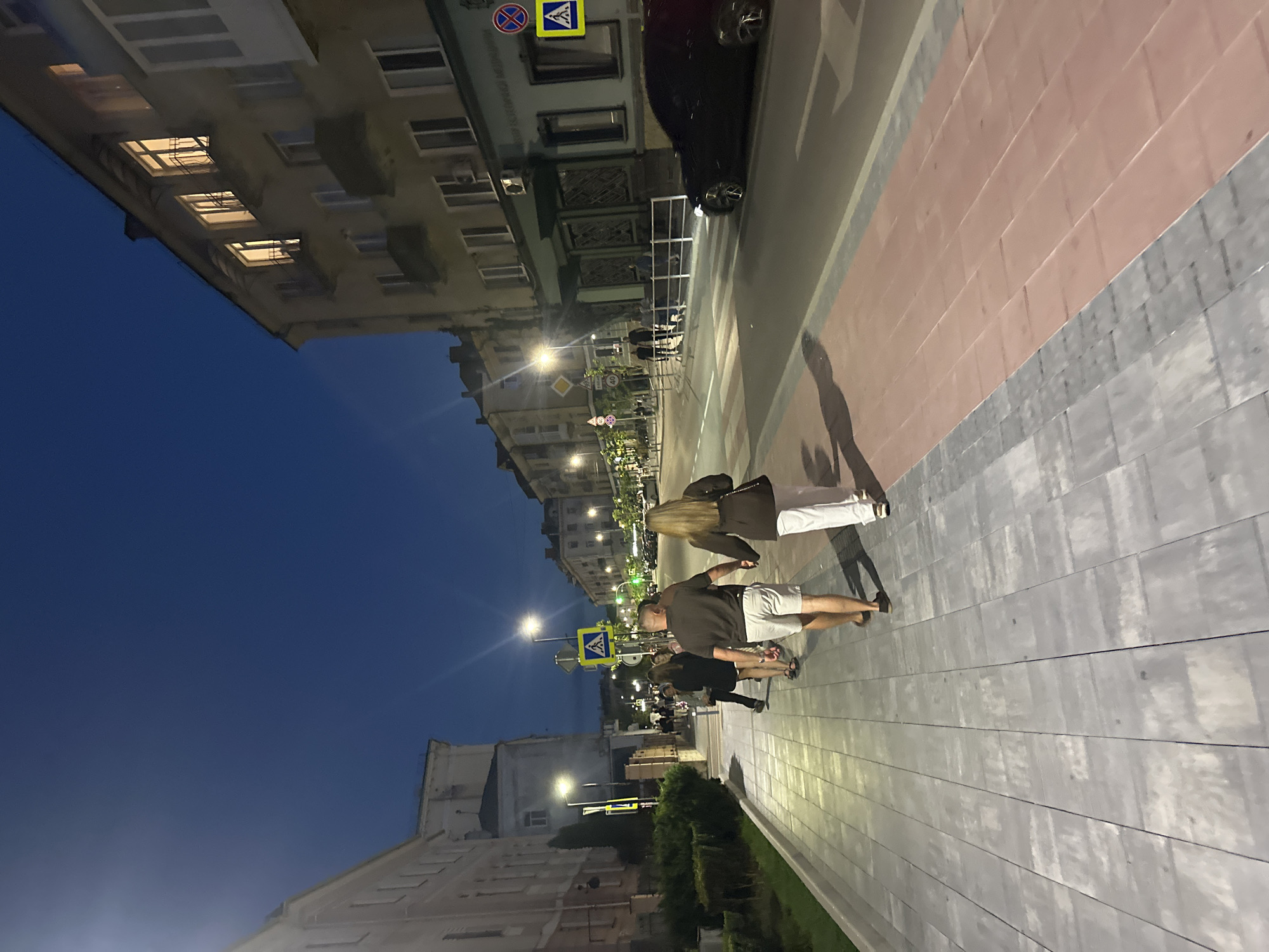 The image depicts a nighttime scene on a city street in Ternopil, Ukraine, likely on one of the main avenues or boulevards, given the presence of multi-story buildings, streetlights, and a crosswalk. A couple is walking hand-in-hand on a paved pedestrian walkway, illuminated by streetlights and the lights emanating from the windows of the buildings. The man is wearing a dark t-shirt and light-colored shorts, while the woman is wearing a dark jacket and white pants. Behind them, other pedestrians are visible, also walking along the sidewalk. A black car is crossing a crosswalk in the street. The buildings on the right side of the street are residential or commercial, with windows lit, suggesting occupancy. The sky is dark blue, indicating it is nighttime. The street is paved, with distinct lanes and markings. Traffic signs are visible, including a pedestrian crossing sign. The overall atmosphere is that of a typical evening in an urban environment.
