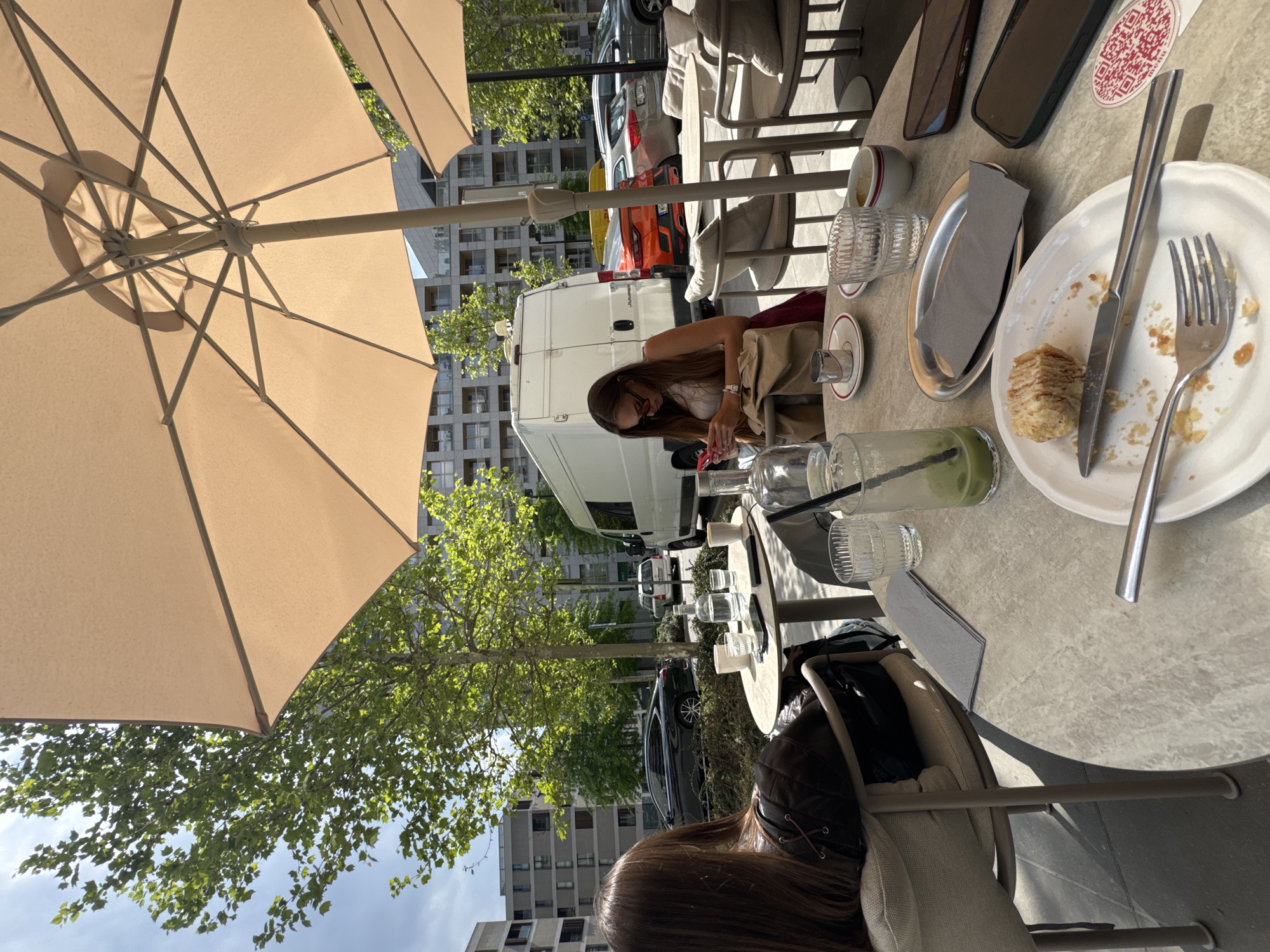 Here is a detailed description of the image:
The image captures an outdoor cafe scene in the city of Unknown, Unknown. Two women are seated at separate tables. One woman is seen in profile, wearing sunglasses, a white top, and tan pants, seemingly occupied with her phone. The other woman is seen from behind, with long brown hair and a jacket draped over her chair.
The tables are set with various items indicative of a meal. One table has a plate with a partially eaten pastry, a knife, a fork, a napkin, a glass of green liquid, a cup, a QR code card, and two mobile phones. The other table has a similar setup. The scene is shaded by large beige umbrellas, and there are glimpses of trees and buildings in the background. Several cars, including a white van, are parked in the street. The overall atmosphere suggests a relaxed, sunny day at a cafe.