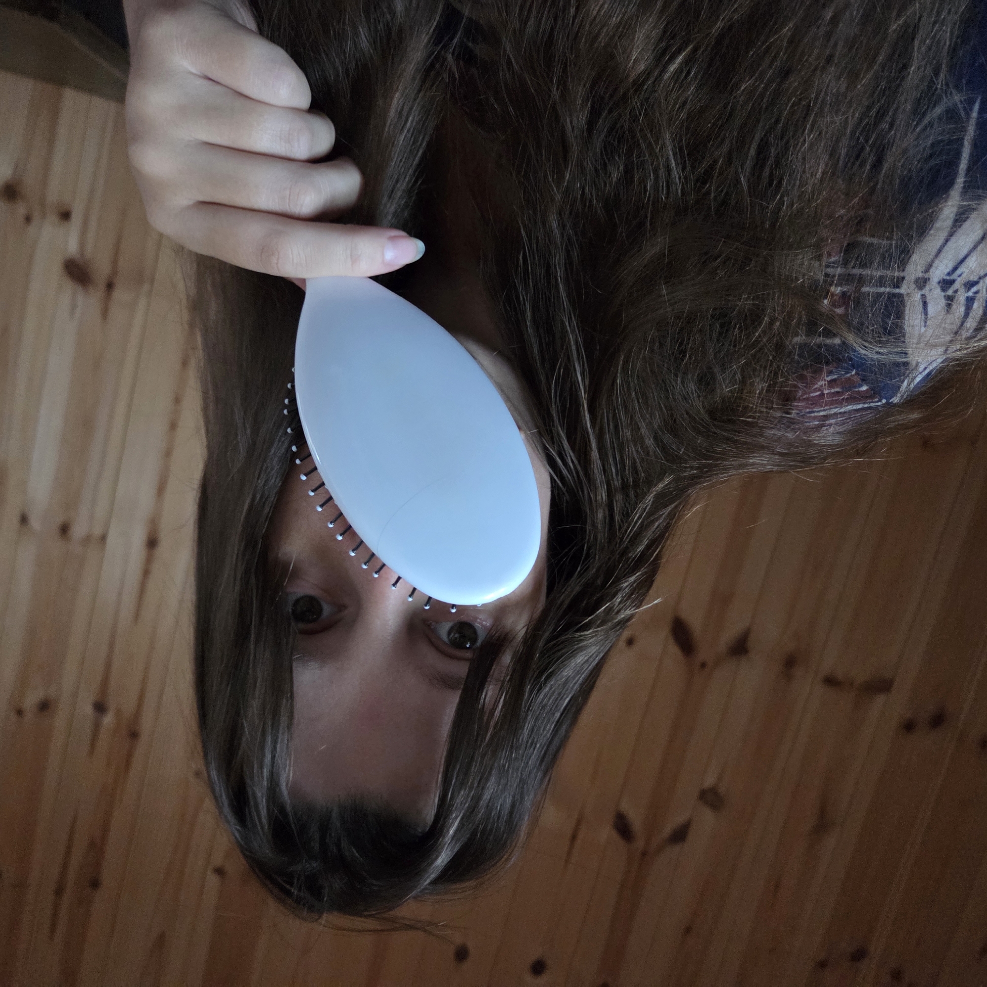 In an indoor setting, with a backdrop of wooden paneled walls, a person's face is partially obscured by a white oval-shaped hairbrush held in their right hand. The brush is positioned in front of the person's nose and mouth, with its bristles visible. The person has long, dark brown hair that cascades around their face and shoulders. Their eyes, visible above the brush, are dark brown. The person is wearing a dark-colored shirt with a pattern of blue, white, and red. The image is cropped to focus on the upper body of the individual and the hairbrush. The lighting suggests an indoor environment, possibly a room with wooden walls. There is no specific information about the location being in Dunnae-myeon, South Korea.