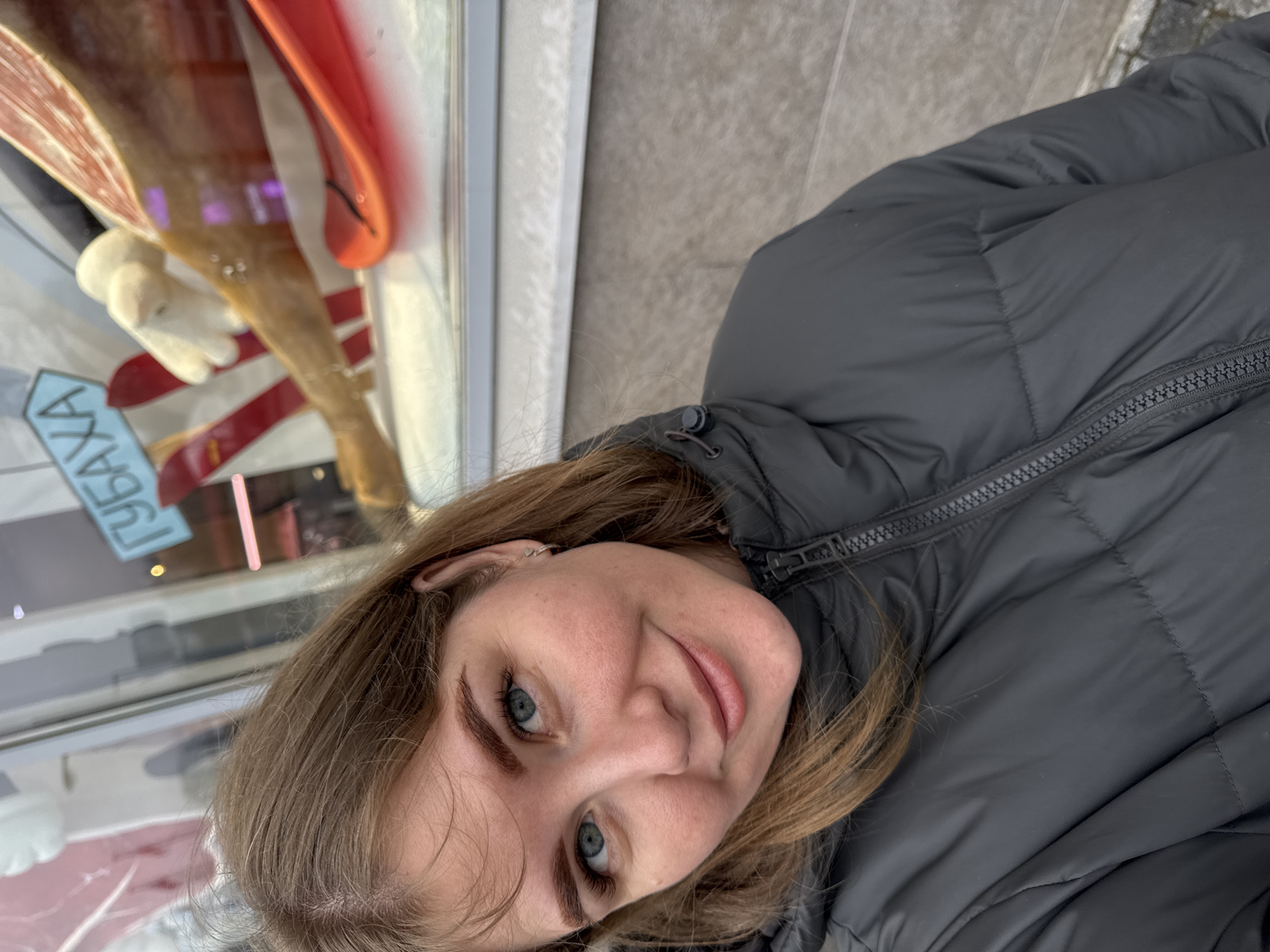 A young woman with fair skin and blue eyes is positioned in the foreground, wearing a dark gray puffer jacket. She is looking directly into the camera with a slight smile. Behind her and slightly to the left is a store window containing what appears to be a jamón display. The signage in the window includes Cyrillic characters. Given that the location is Perm, Russia, and the presence of jamón, it suggests the store could be a deli or specialty food shop selling Spanish cuisine. The background also includes a gray stone surface, possibly a sidewalk.