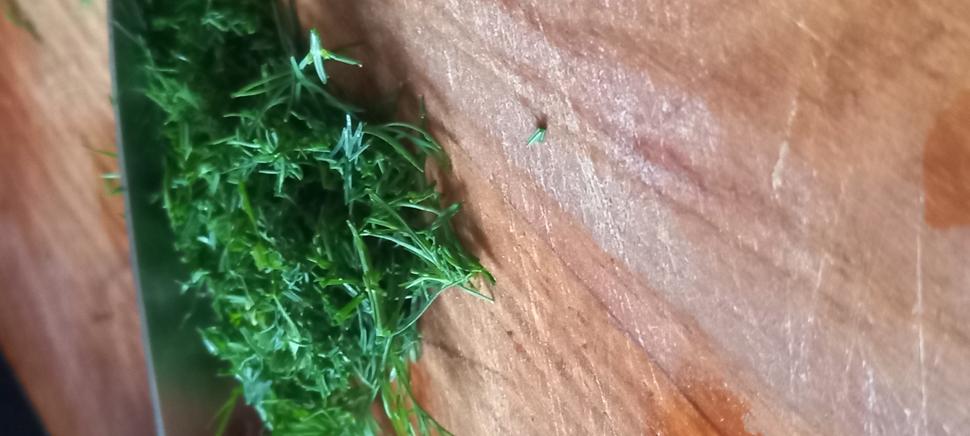 A close-up, high-angle view captures a pile of freshly chopped green herbs, identified as dill, resting on a wooden cutting board. The bulk of the chopped herbs occupies the upper-left and central regions of the frame. The dull silver blade of a knife is partially visible horizontally across the upper portion of the image, positioned above the herbs, indicating recent preparation. The wooden surface of the cutting board exhibits a light brown hue with visible grain patterns and scattered minor scratches. A single, small, detached piece of a chopped herb is present on the bare wooden surface towards the mid-right. No individuals, additional objects, or further environmental context beyond the cutting surface are discernible. This scene is located in Kryvyi Rih, Ukraine.