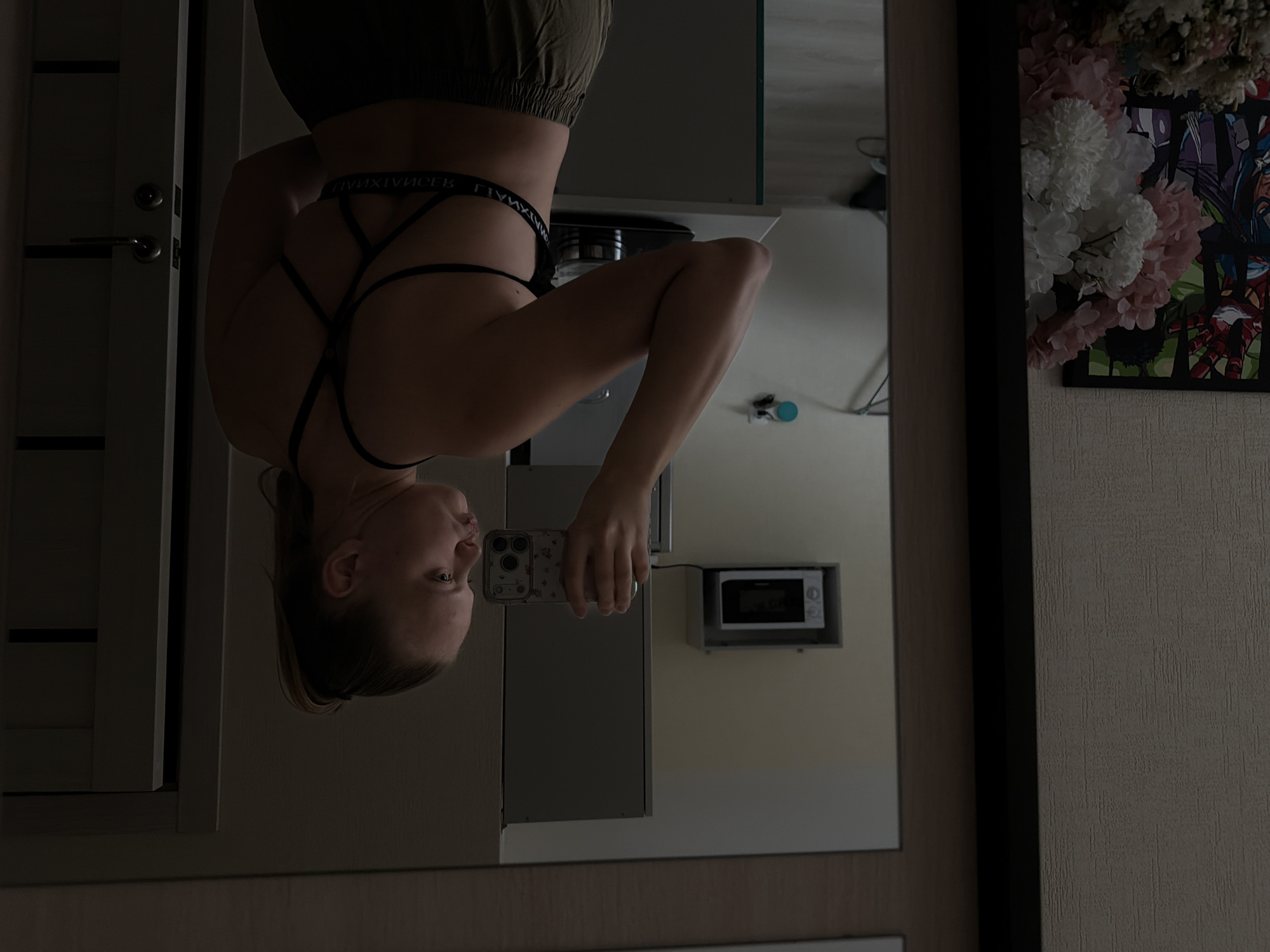 A woman is taking a selfie in a mirror. She is wearing a sports bra and pants. The background includes a door, a microwave, and some decorative flowers and a picture on the wall. The location is Izhevsk, Russia.