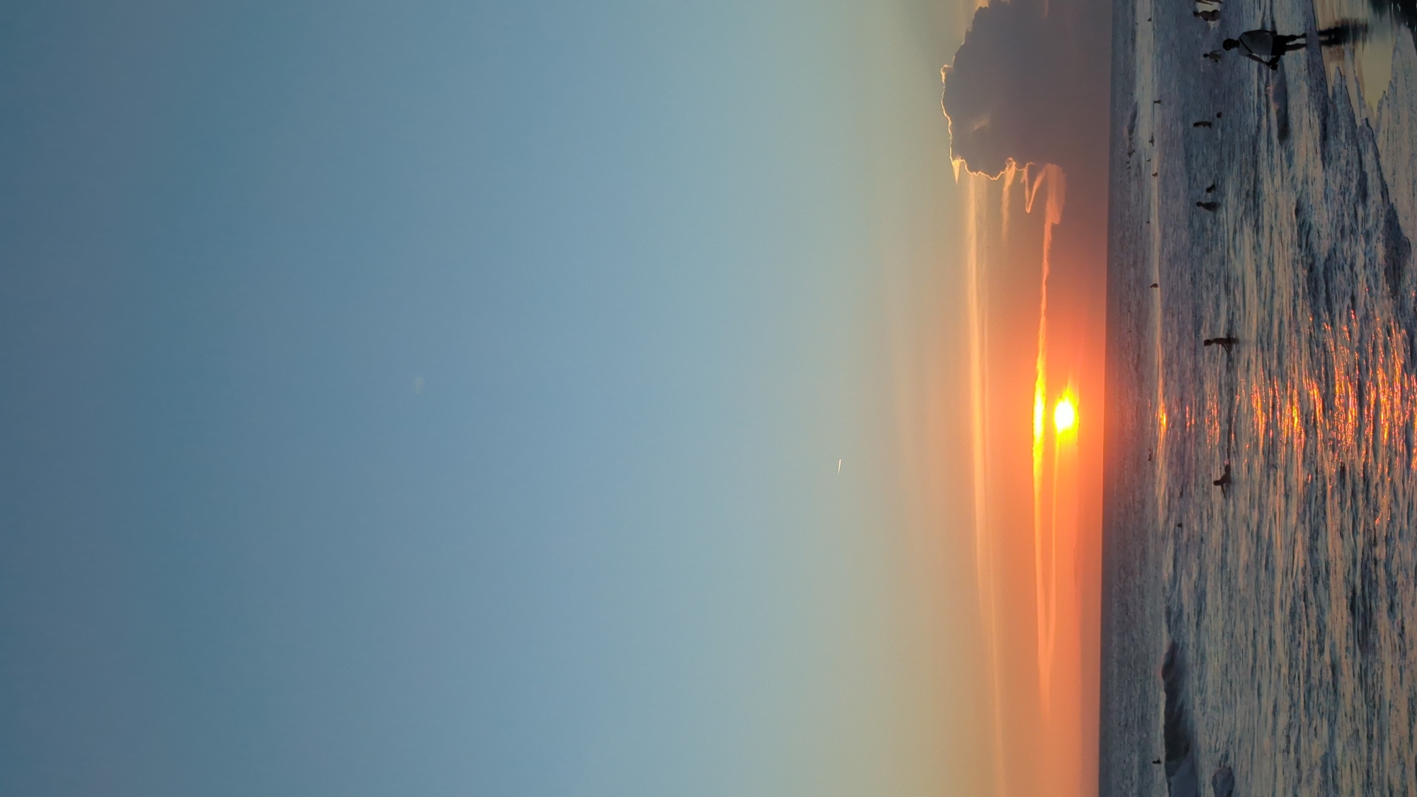 The image captures a sunset scene at a beach, possibly in North Kuta, Indonesia, given the composition. The sky transitions from a clear blue to a warm orange near the horizon, where the sun is setting. A solitary cloud with bright edges sits above the sun. The ocean reflects the colors of the sunset. Several people are visible in the water, appearing as silhouettes, with one individual standing near the shore. The waves are reflecting the bright sunlight, creating a shimmering effect on the water's surface.