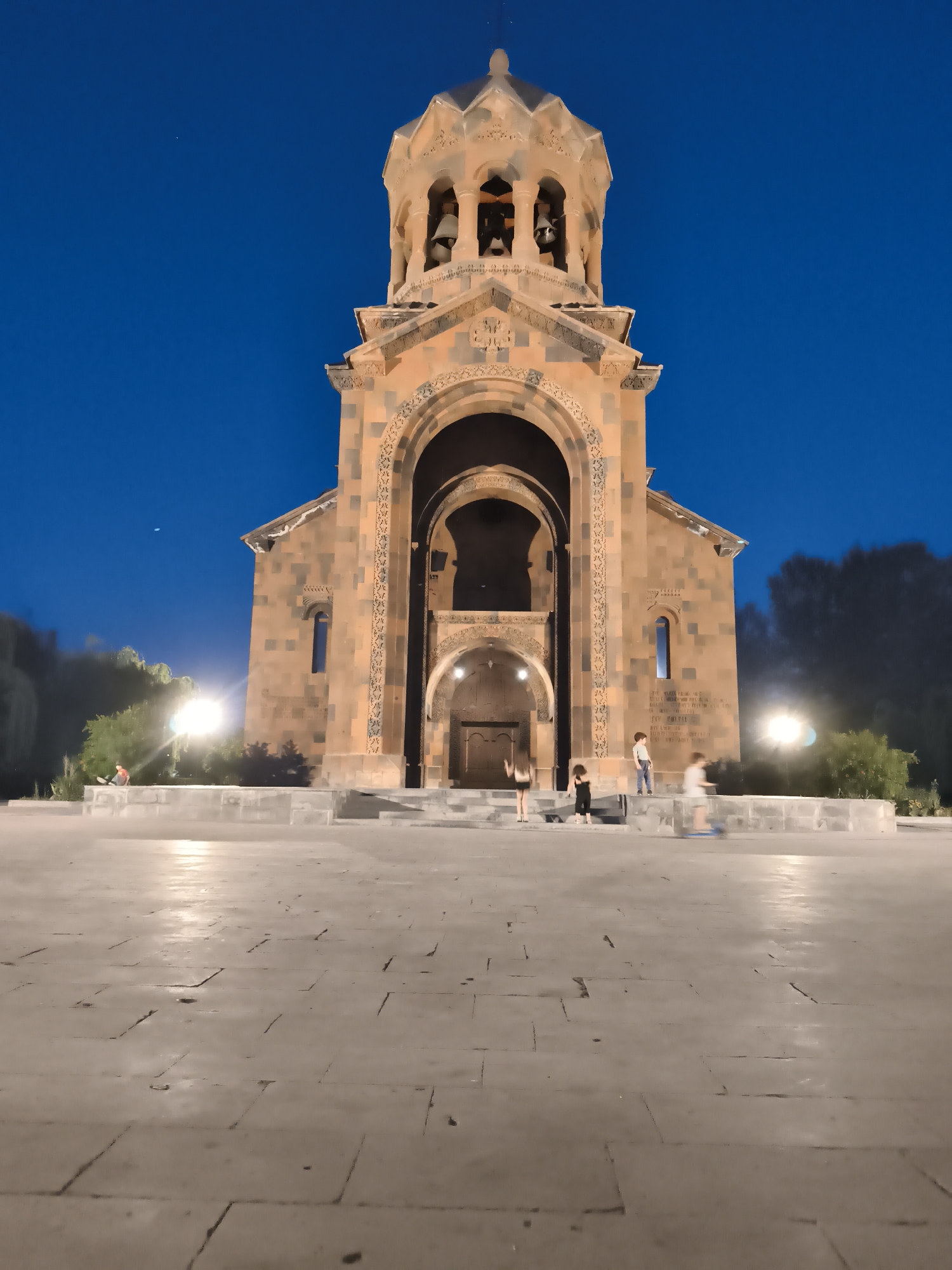 A wide-angle, eye-level shot captures a religious building with a bell tower at dusk in an outdoor, urban setting. The building, constructed of tan and dark orange stone, features intricate carvings around its arched entrance. The bell tower, topped with a multi-faceted dome, has arched openings revealing bells. The ground in front of the building is paved with large, light-colored stones. Several small figures, appearing to be children, are present on the steps and in the plaza. One child is on a scooter, blurred due to motion. Trees and streetlights are visible in the background. The sky is a deep, clear blue.