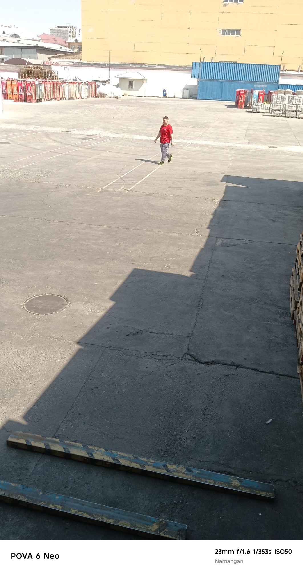 An outdoor, overhead shot captures a lone young boy wearing a red t-shirt and grey shorts, walking away from the camera in a wide, paved, outdoor area. The ground is concrete with visible cracks and painted lines. To the left of the boy, a circular metal manhole cover is set into the pavement. In the background, a long, tan-colored building with visible windows stretches across the frame, with a row of blue shipping containers behind it. A small white building with a dark roof is situated near the center of the frame. The overall scene is likely a port or industrial area, with stacks of pallets and containers visible. The image is tagged with "23mm f/1.6 1/353s ISO50" and "Namangan," indicating it was taken in Namangan City, Uzbekistan.