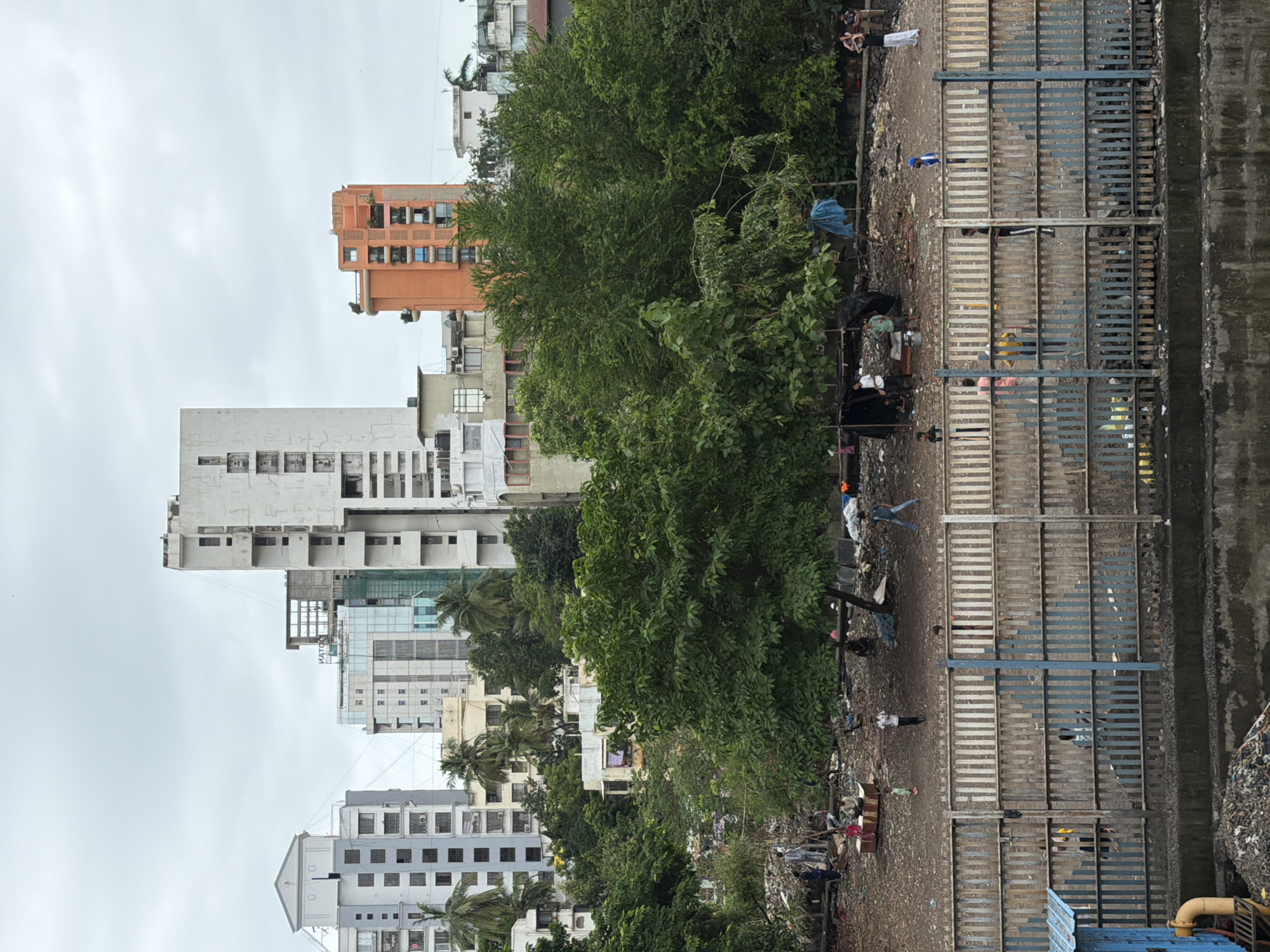 The image shows a slum area adjacent to a fence and buildings in Mumbai, India. In the background, there are several multi-story buildings, including a tall white building with a distinctive geometric design and an orange building with balconies. In the foreground, a densely wooded area with large green trees separates the slum from a fence. The slum itself appears to be an open area with scattered debris, makeshift shelters, and a few people. Some individuals are seen sitting, standing, or walking around. The fence in the middle ground is made of vertical metal bars and appears to be a barrier between the slum and possibly a construction site or a different part of the city. The sky is overcast, suggesting cloudy weather. The scene depicts a stark contrast between urban development and informal settlements in Mumbai.