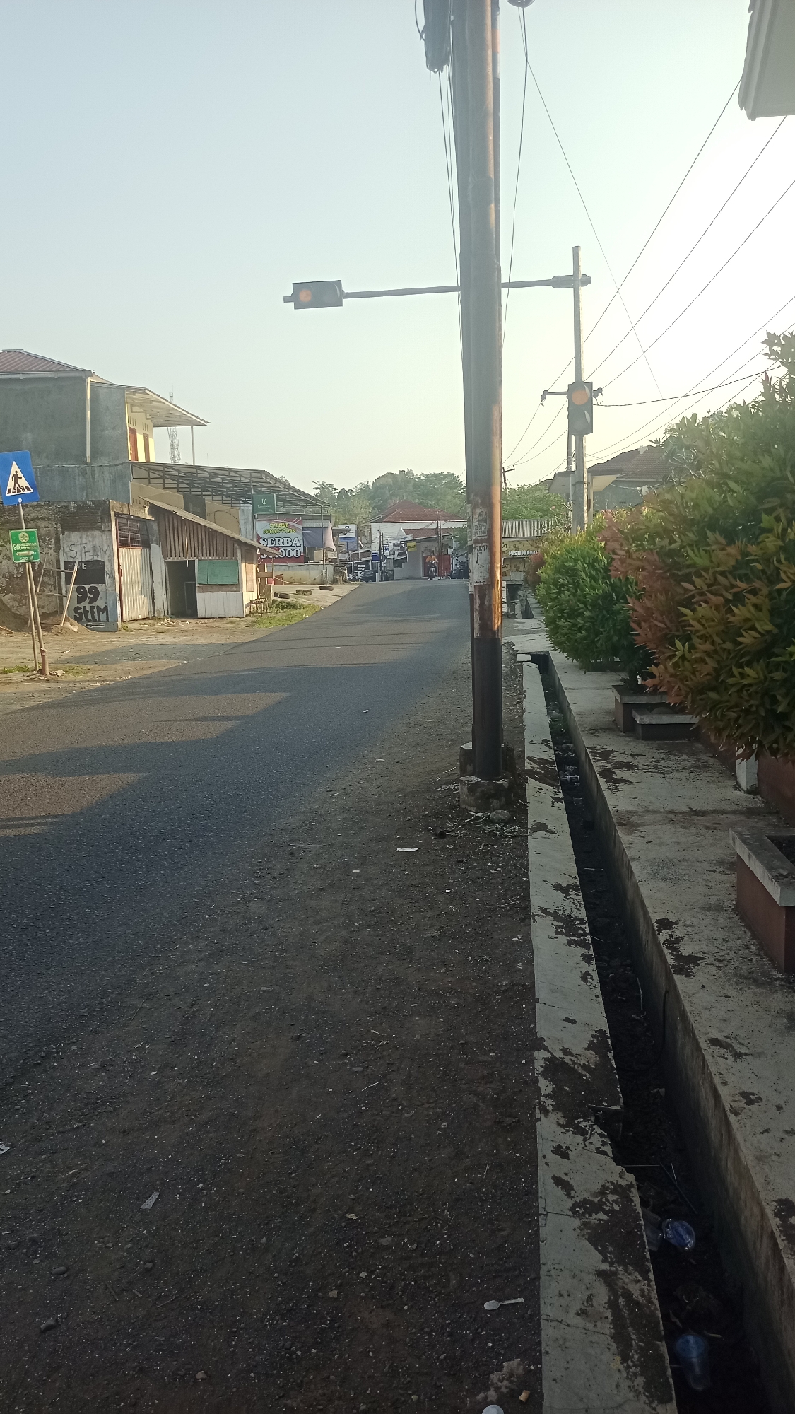 The image depicts a street scene in Karyamandala, Indonesia, under a clear sky with a soft, warm light, suggesting either early morning or late afternoon. The road surface is asphalt, with a textured dirt shoulder on the left and a concrete drainage ditch on the right.
On the left side of the road, there are several buildings. The closest building is a single-story structure with a light-colored wall and a corrugated metal roof supported by a dark frame. A pedestrian crossing sign, a blue diamond with a white silhouette of a person walking, is mounted on a pole. Adjacent to this is a smaller, greenish-blue structure with an open entrance. Further down the street, more buildings are visible, some with awnings and shop signs, including one with "SERBA 9.000" printed on it. Graffiti marks the wall of the closest building.
A tall, weathered wooden utility pole stands prominently on the right side of the image, supporting multiple power lines. Attached to it are two traffic lights. The one on the left, over the road, shows a yellow light. Another traffic light, further down the street on a separate pole, also displays a yellow light.
The right side of the scene features a concrete drainage channel that appears to be filled with some debris, including plastic bottles and other litter. Beyond the drainage channel, there are manicured bushes with red and green foliage, planted in raised beds. In the background, residential buildings with tiled roofs are visible, nestled amongst trees.
A few vehicles are present on the road in the distance, including what appears to be a motorcycle and a small car. No people are in the immediate foreground, but figures can be inferred in the distant vehicles and shops. The overall impression is of a quiet, developing street on the outskirts of a town.