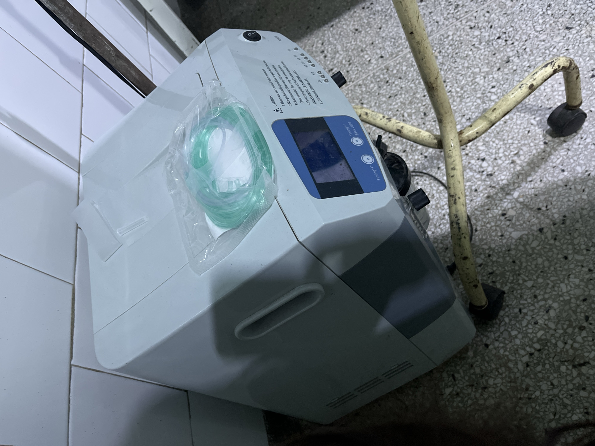 The image shows a medical oxygen concentrator in a hospital or clinic setting. The concentrator is white and gray, with a blue-lit display screen and a clear plastic bag containing a green oxygen tube attached to it. There are caution labels and operation instructions printed on the top of the machine. It sits near a white-tiled wall and a mobile stand, likely for transporting the device. The floor is tiled with a speckled pattern. There is no mention of the city of Juan Griego, Venezuela, so I cannot provide any details on that.
