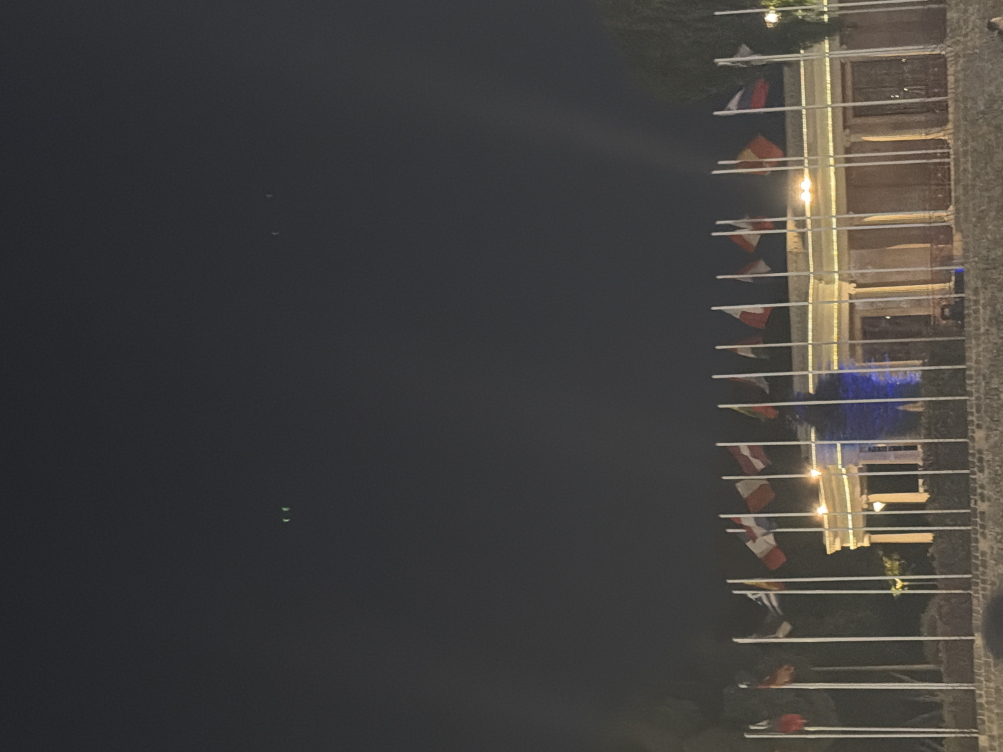 The image depicts an outdoor nighttime scene with a building illuminated by lights. A series of flags are displayed on poles extending horizontally from the building's facade, creating a decorative line. Some of the flags are visible as red, white, and yellow, suggesting they may be national or organizational flags. The building appears to have a traditional architectural style, with stone elements and multiple windows. The overall atmosphere is festive, possibly indicating an event or celebration. The sky is dark, implying it is nighttime, and the lighting from the building casts a warm glow on the surroundings. There are no visible people or text in the image. The location context is Nicosia, Cyprus.