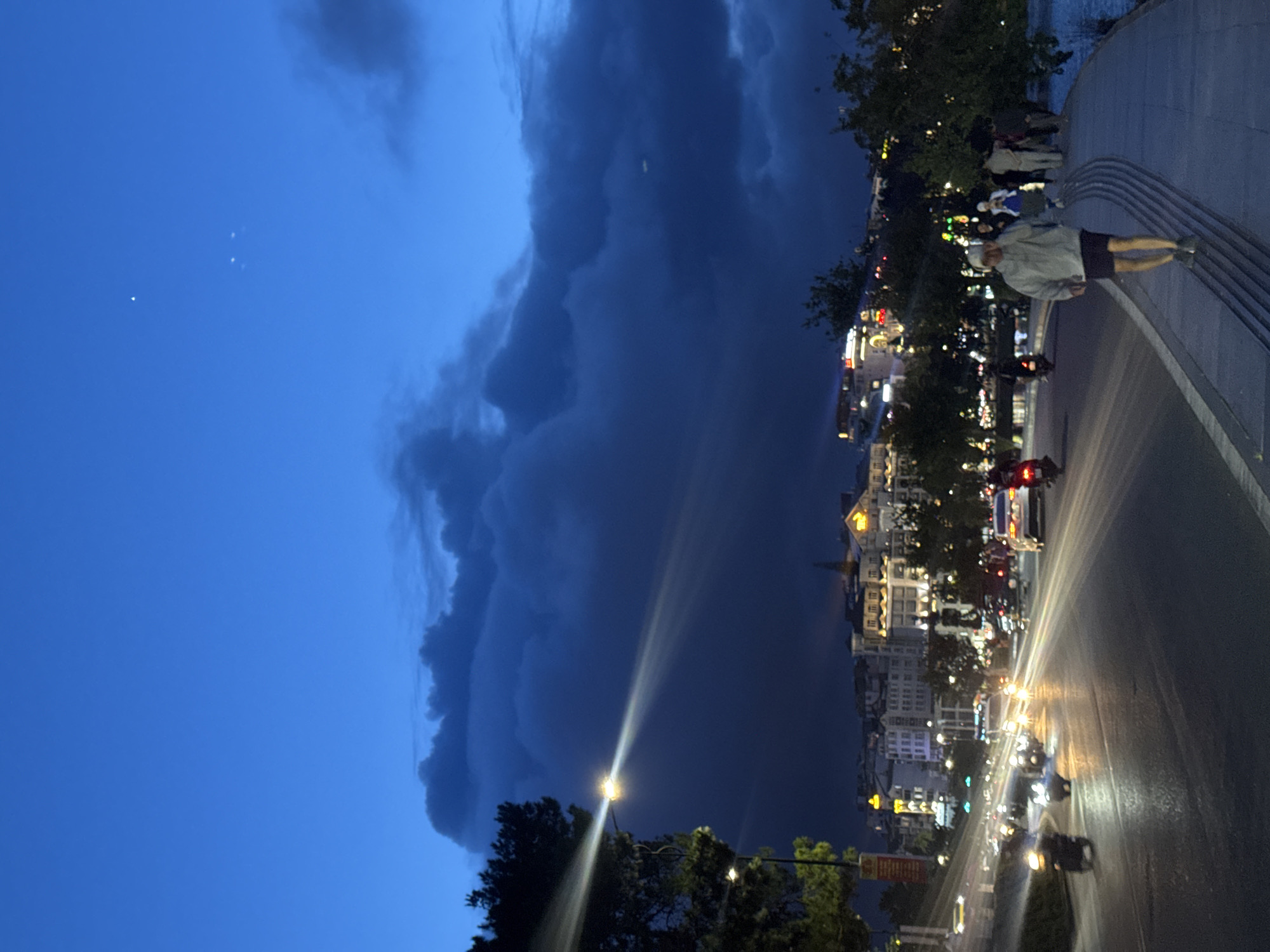 This photograph captures a dramatic urban scene in Da Lat, Vietnam, at twilight or early night. The upper portion of the image features a striking contrast in the sky: a vast expanse of clear, deep blue on the left, dotted with a few faint specks of light, gives way to an enormous, dark, and heavy cloud formation that dominates the right half of the sky, suggesting an impending or recent storm.
Below this dramatic sky, a vibrant city street stretches across the lower part of the frame. The road appears wet, reflecting the numerous bright headlights and taillights of vehicles, predominantly motorbikes, creating elongated streaks of light as they move. Several cars are also visible in the traffic flow. Buildings line the street, their multiple stories illuminated by internal lights and external signage, adding to the city's glow. A strong, radiant beam from a prominent streetlight slices across the lower left quadrant of the image, likely caused by lens flare.
Parallel to the street, on the far right, is a wide promenade or elevated walkway. Several people are present, including one individual in the immediate foreground, wearing a light hoodie and dark shorts, standing on steps and looking towards the bustling street. Others are seen further back on the walkway. Dark-leaved trees are interspersed among the buildings and along the promenade, providing natural elements to the urban landscape. The overall atmosphere is dynamic and atmospheric, blending the activity of the city with the imposing natural weather elements.