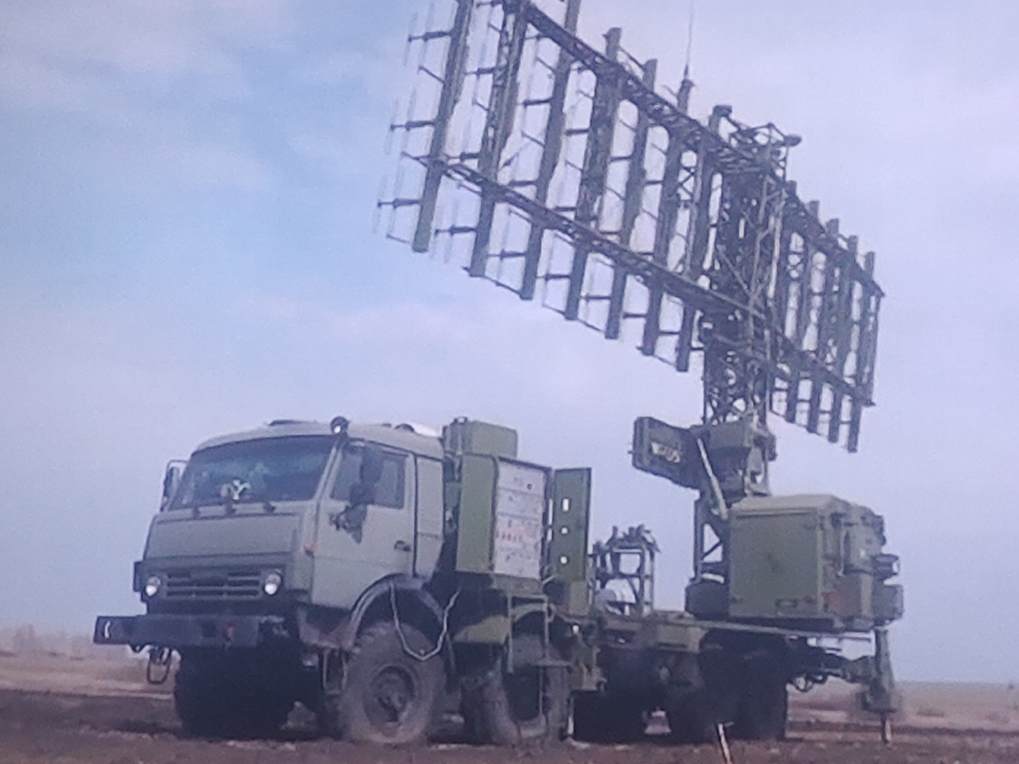 A military radar system is deployed in a field under a hazy sky. The radar array, a large, lattice-like structure with multiple horizontal panels, is mounted on a vehicle. The vehicle itself is a heavy-duty, camouflaged truck with a large cab and multiple axles. The truck is parked on a dirt or gravel surface, with some sparse vegetation visible in the background. The overall scene suggests a mobile military operation. The image does not depict the city of Molenbeek-Saint-Jean - Sint-Jans-Molenbeek, Belgium.