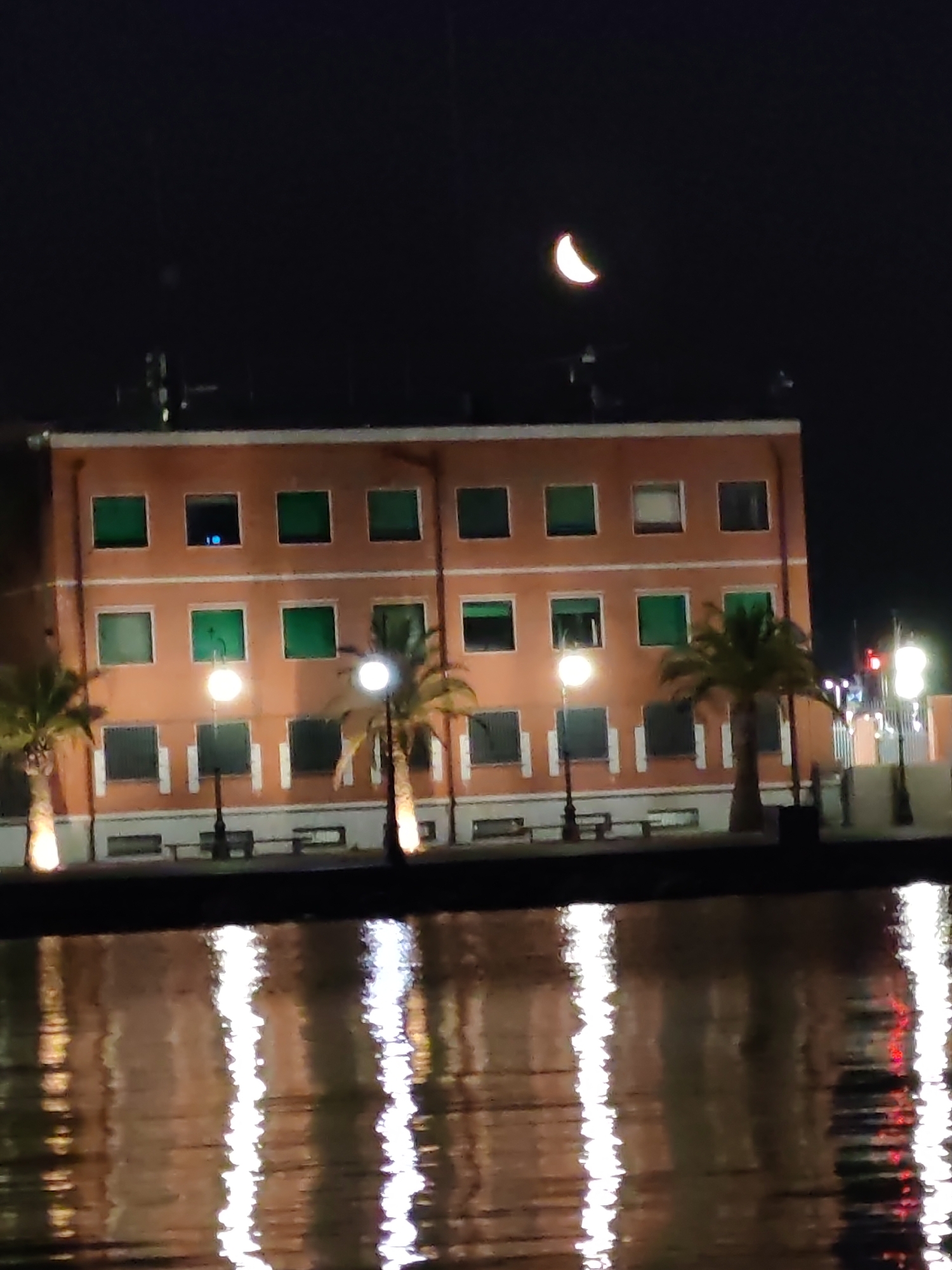 At night, a wide shot captures a building in the city of La Spezia, Italy, which appears to be a three-story structure with a terracotta-colored facade. The building features numerous square windows with green shutters, some of which are closed while others are open, revealing interior lights. A crescent moon is visible in the dark night sky above the building. In front of the building, there is a body of water that reflects the lights of the building and streetlights, creating shimmering patterns. Several palm trees are planted along the waterfront. Streetlights and benches are also present along the building's front. The overall scene depicts a peaceful night view of a waterfront area in La Spezia.