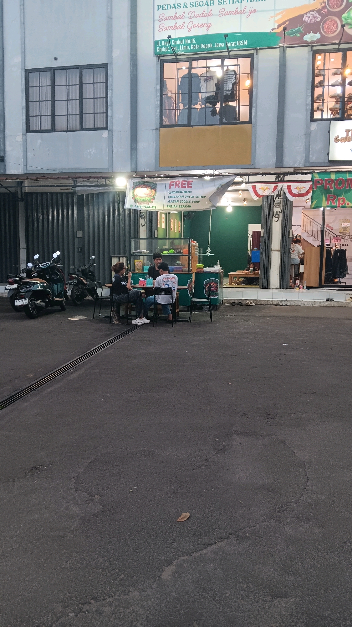 The image shows a street-level view of a restaurant or food stall in Depok, Indonesia. The restaurant is situated in a building with a grey facade and dark-framed windows. A banner promoting "FREE CONDIMENTS/MENU" is displayed above the entrance, with a phone number visible. Inside the establishment, a green wall is visible, along with tables and chairs. Three individuals are seated at a table outside the restaurant, appearing to be customers or staff. Two motorcycles are parked to the left of the scene. The ground in front of the establishment is asphalt, with some visible wear and tear. The overall lighting suggests it is dusk or early evening.
