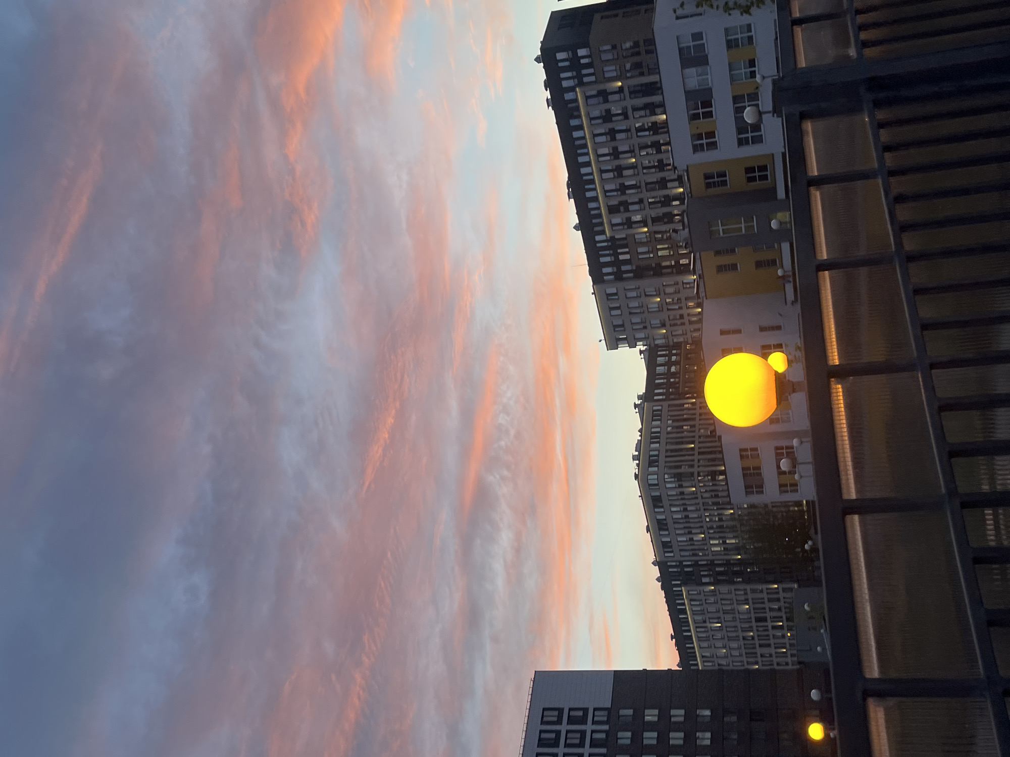 The image shows a view of the sky at dusk in Saint Petersburg, Russia. The sky is filled with wispy clouds tinged with pink and orange from the setting sun. In the foreground are residential buildings of varying heights and colors, mostly gray and white. Streetlights shaped like yellow globes are visible near the buildings. A dark metal fence or balcony railing occupies the right side of the frame.