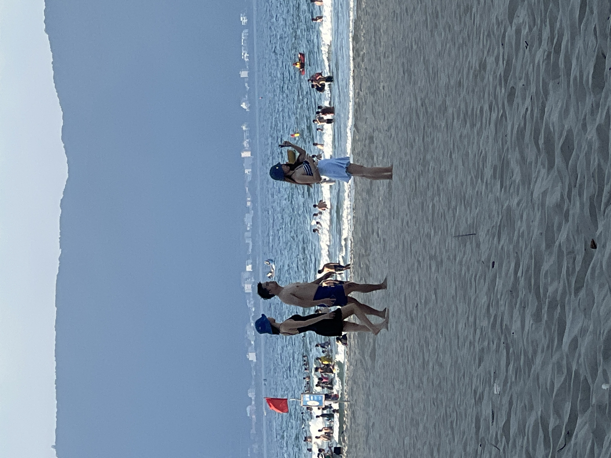 The image depicts a vibrant scene at a crowded beach in Đà Nẵng, Vietnam. The setting is a sandy beach with the ocean in the background, suggesting a warm, sunny day. The water is a brilliant blue, with gentle waves lapping the shore. Numerous people are visible enjoying the beach, some wading in the water, others sunbathing, and a few are playing closer to the shore.
In the foreground, three individuals are the primary focus. Two people, a man and a woman, are standing close together. The man is shirtless, wearing blue swim shorts, while the woman is in a black swimsuit and a blue cap. Slightly further back, another person, possibly a woman, is standing alone and appears to be taking a selfie with her phone. She is wearing a blue and white striped top and light blue shorts, also with a blue cap. The group appears relaxed and engaged in typical beach activities.
Further out in the water, several other people are visible, some on what appears to be a small inflatable raft. The beach itself is sandy, with visible ripples and patterns in the sand, indicating it has been recently raked or shaped by the wind and waves. In the distance, a line of buildings suggests an urban area bordering the beach. A red flag is prominently displayed on a pole, which might indicate a specific safety warning or zone. The overall impression is one of a lively summer day at a popular tourist destination. The lighting suggests it is daytime, likely afternoon given the angle of any shadows.
