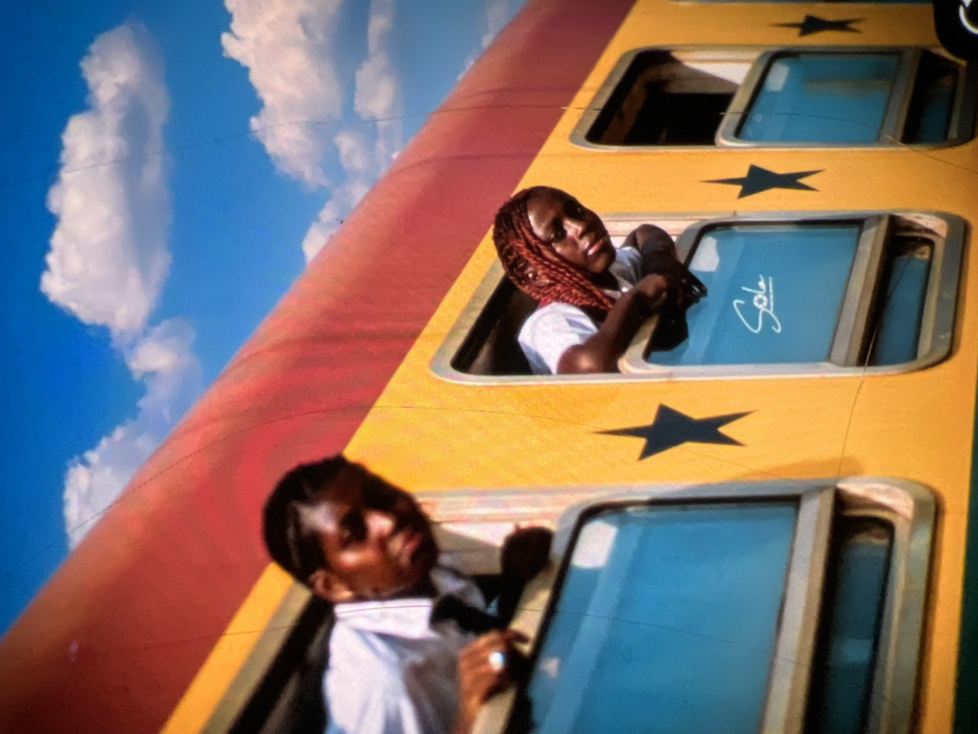 In a medium shot, two young Black individuals lean out of the windows of a bright yellow and red train car under a vibrant blue sky with white clouds. The person on the left, wearing a white shirt and a dark tie, looks forward and slightly down, with their face and upper body visible. They are wearing a ring on their left index finger. The person on the right, with braided red hair, is also looking forward and down, their arms resting on the windowsill. Their white t-shirt is visible. The train car has dark blue windows, some of which bear a black star graphic. The window on the right displays the word "Sole" written in cursive white lettering. The scene appears to be set in Lagos, Nigeria, possibly indicating travel or a journey. The overall impression is one of youthful energy and movement.