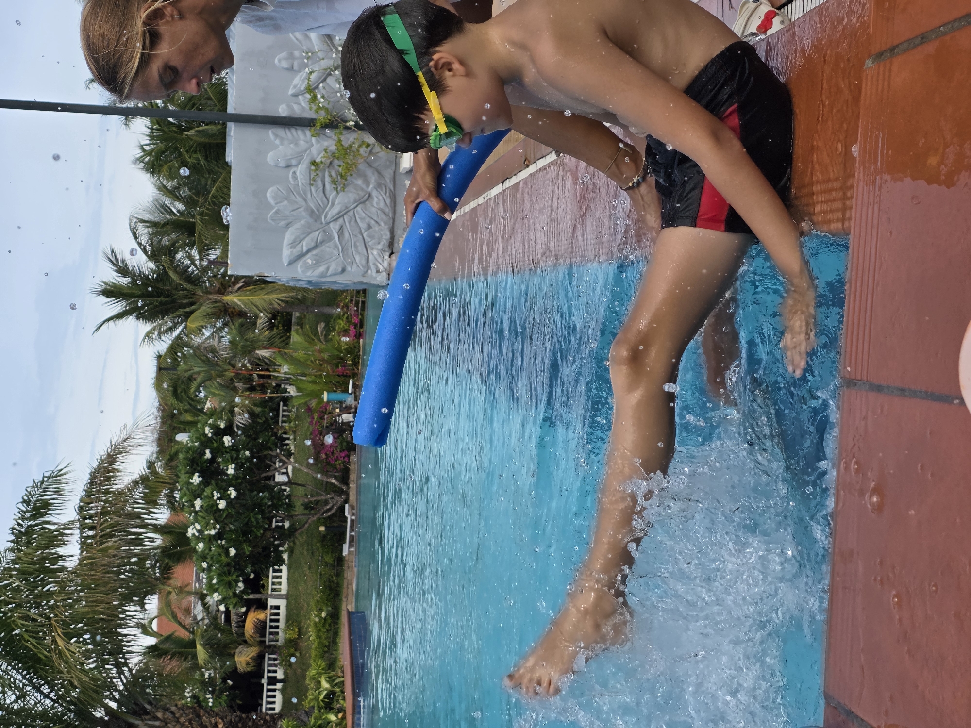 A boy wearing goggles and swim trunks is entering a pool in Phu Thuy Ward, Vietnam. He is holding a blue pool noodle. A woman is nearby. Palm trees and buildings are in the background. Water is splashing around the boy's legs as he enters the pool.