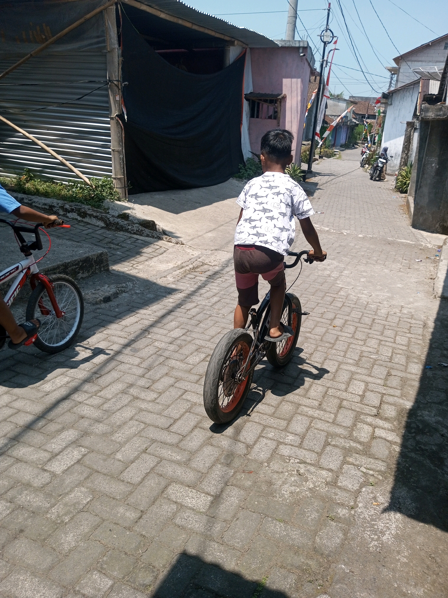 A young boy is riding a bicycle down a paved alleyway in Andongsili, Indonesia. He is wearing a white t-shirt with a pattern of sharks and dark shorts with pink trim. The alley is lined with houses and buildings, some of which appear to be under construction or in disrepair. There are also several motorcycles parked along the side of the alley, and Indonesian flags are visible. The sun is shining, casting shadows on the ground. Another bicycle is partially visible in the foreground to the left.