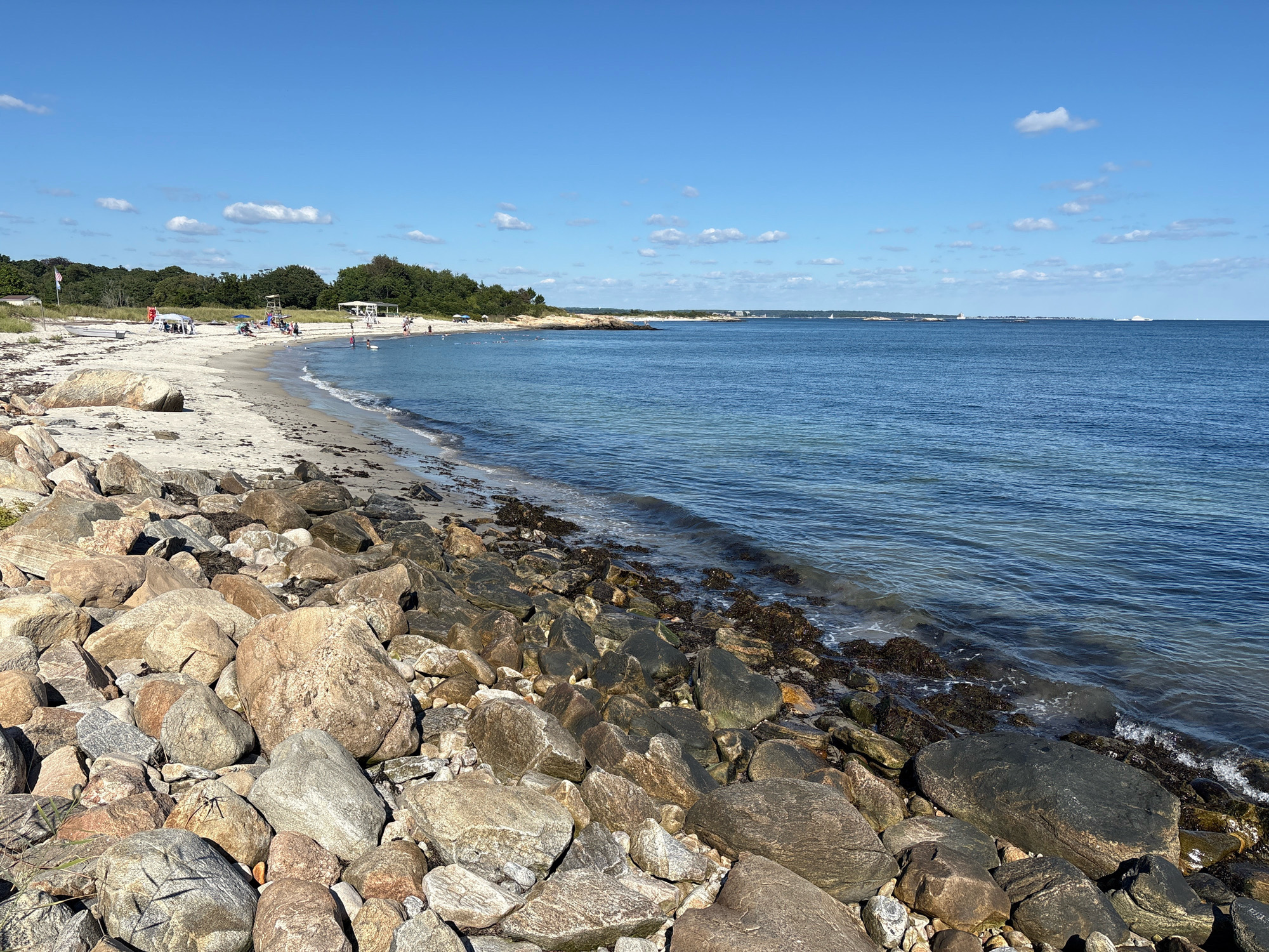 The image depicts a scenic view of Waterford, Connecticut, USA, specifically at Ocean Beach Park. The foreground is dominated by a rocky shoreline with numerous large boulders of varying shades of grey, brown, and tan. Beyond the rocks, a sandy beach stretches into the middle ground, dotted with a few people and beach umbrellas. The water of Long Island Sound is calm and blue, with gentle waves lapping at the shore. In the distance, lush green trees line the coastline, and several boats are visible on the horizon under a clear blue sky with scattered white clouds. The overall atmosphere is serene and idyllic, suggesting a pleasant day at the beach.