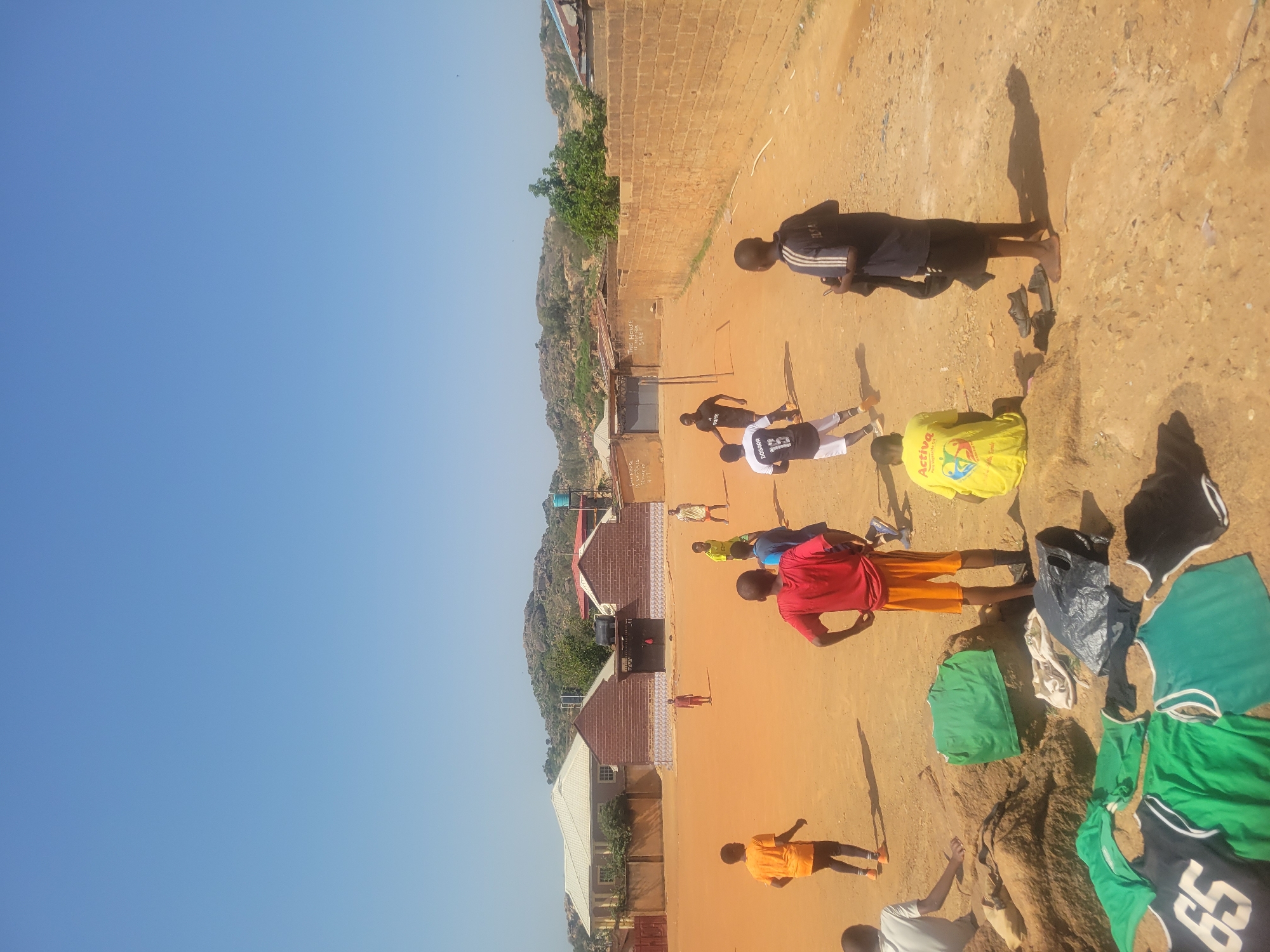 The image shows a dirt field in a residential area of an unknown city. Several people, mostly young males, are standing or lying on the ground. Some wear sports clothing. Clothes, including jerseys and shorts, are scattered on the ground. Buildings and rocky hills are visible in the background. The sky is clear blue.