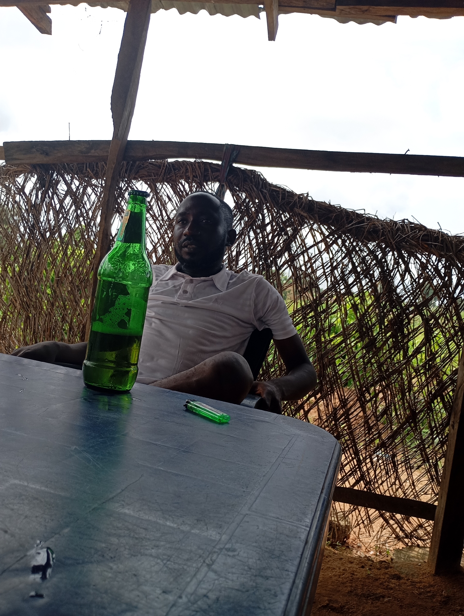 A Black man is seated at a dark blue table under a thatched roof in Shendam, Nigeria. He wears a white polo shirt and has a green bottle of beer in front of him, which is half-full. A green lighter rests on the table to the right of the bottle. The background shows a rustic woven wall with greenery visible beyond it.