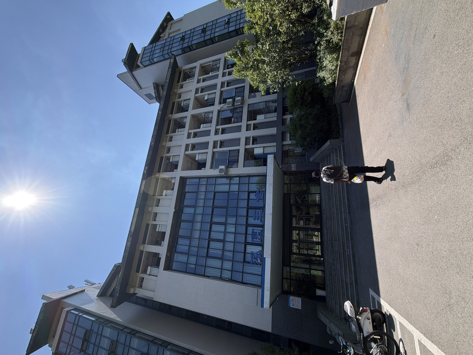 A wide shot captures a man walking away from the camera towards a large, multi-story building with a blue sky and bright sun overhead. The building appears to be made of concrete and glass, with a tiled facade and several balconies. The man is wearing a backpack and carrying a bag. In the foreground, a white scooter is parked on the left side of the frame. The scene suggests an outdoor setting, possibly a university campus or a residential complex, given the architectural style and the presence of a scooter. The image is taken from a low angle, emphasizing the size of the building and the vastness of the sky. The sun is high in the sky, casting strong shadows and indicating it is likely midday. The building has large windows, some of which are open, and air conditioning units are visible on some of the balconies. The entrance to the building features a set of stairs leading up to glass doors. The man is walking on asphalt pavement. Green trees and bushes are visible on the right side of the building. The architectural style of the building, including the rooflines and the decorative elements, suggests it is located in China. Specifically, based on available information, this building is the Sanyuan Dormitory at Southwest University in Jiangjin District, Chongqing, China.